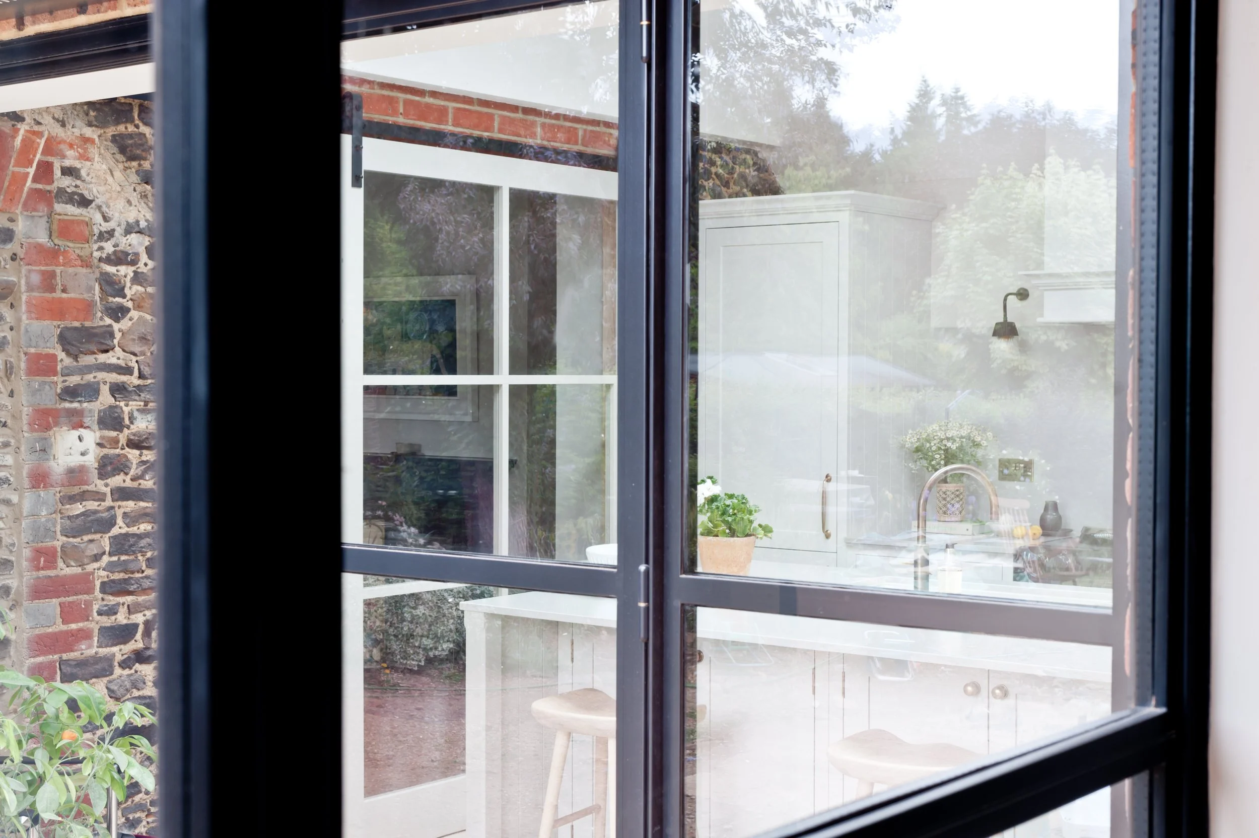 View through a window showing a white outdoor kitchen with a sink and potted plant in a backyard, with a brick wall and trees in the background.