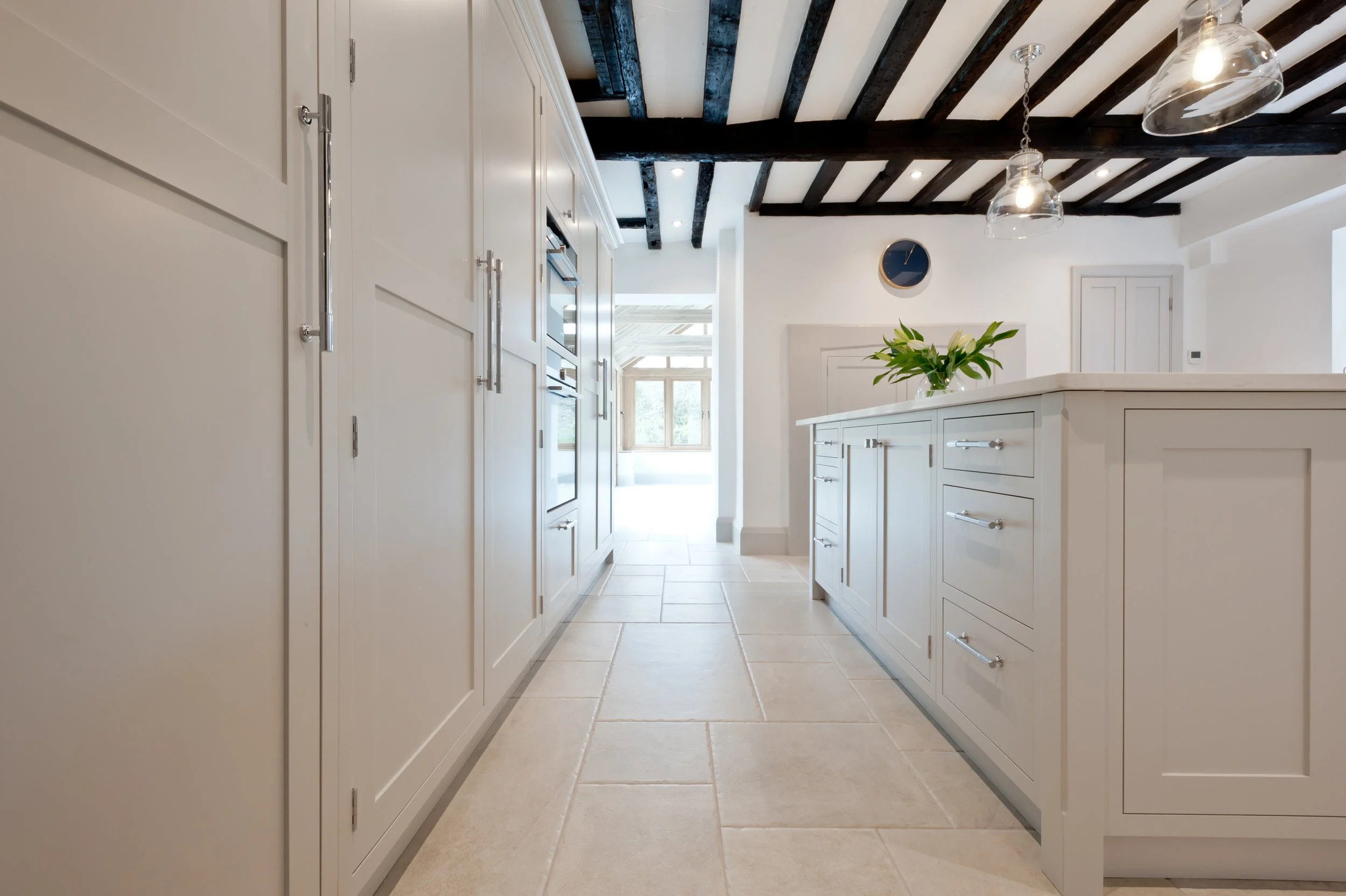 Modern kitchen with white cabinets, a kitchen island with a flower vase, tiled floor, exposed dark wooden ceiling beams, and large windows.