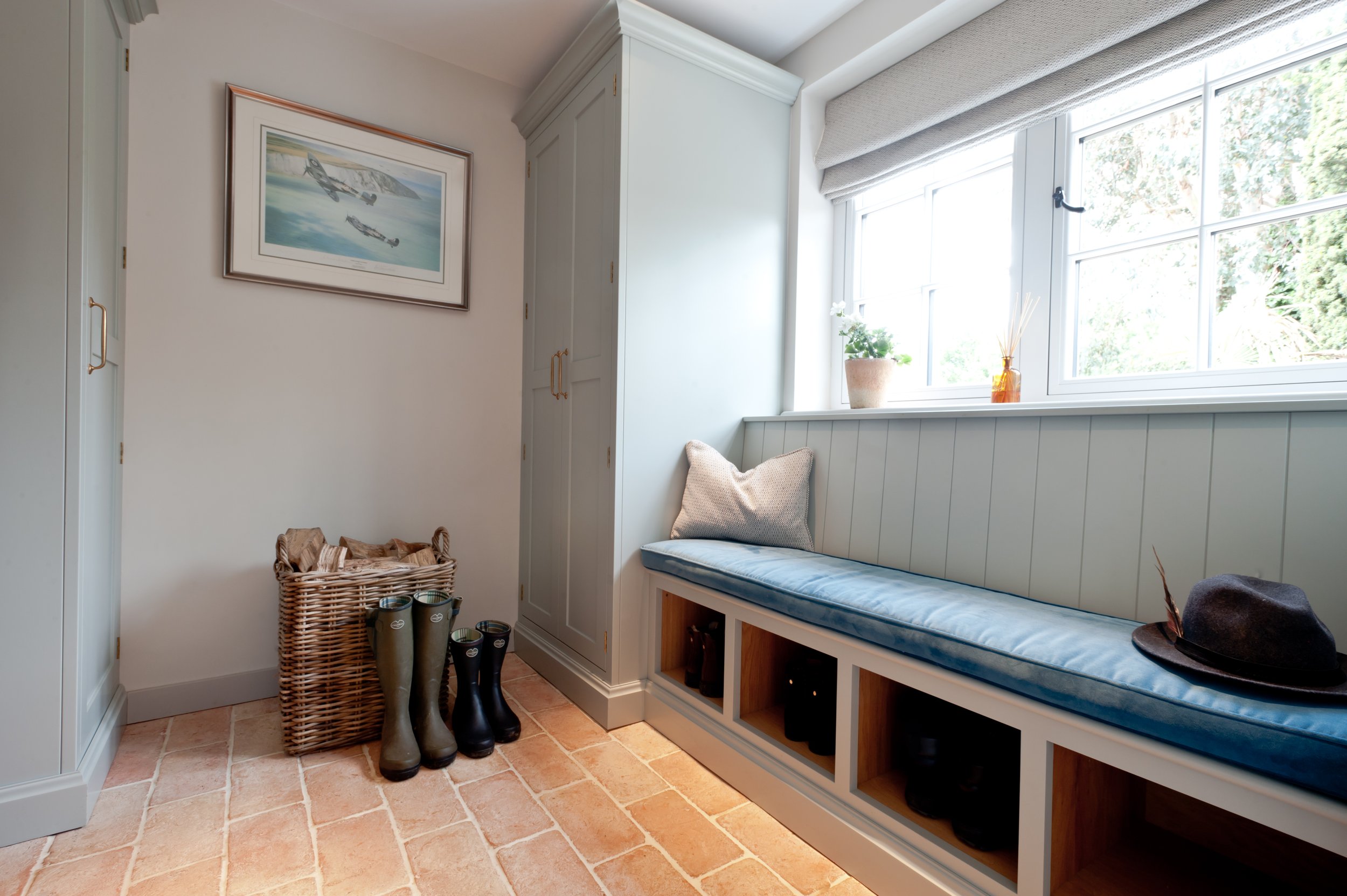 Bespoke boot room cabinetry in soft grey-green with a cushioned window seat, open shoe storage, tall cupboards and terracotta brick flooring.