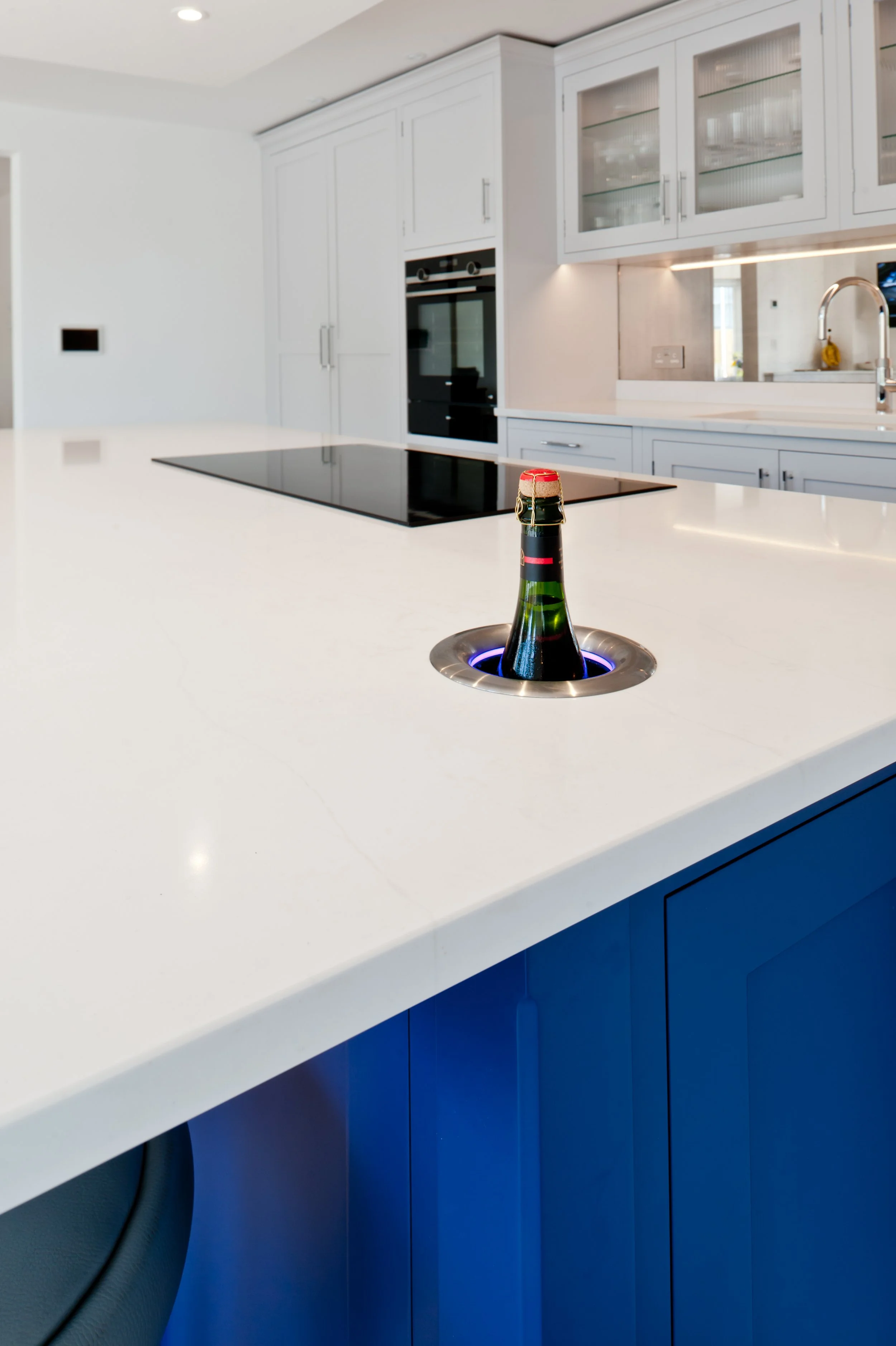 Modern kitchen with a white countertop, blue cabinets, a black cooktop, and a built-in oven. There is a bottle of champagne in an indented opening in the countertop.
