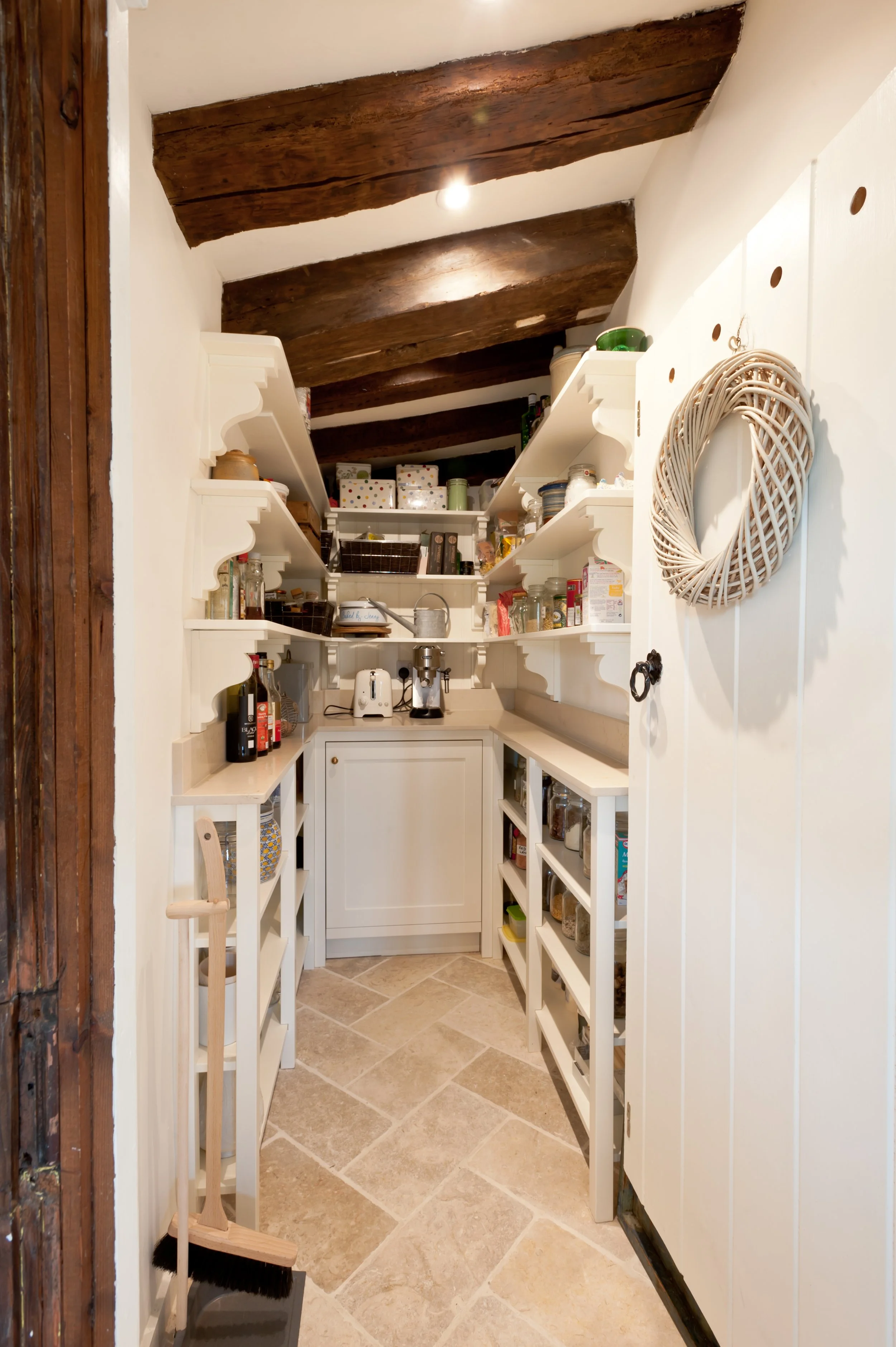 A small pantry with white shelving filled with jars, boxes, and kitchen essentials, cream-colored tile floor, exposed dark wooden ceiling beams, and a white cabinet with counter space. There is a brush, dustpan, and broom leaning against the wall.