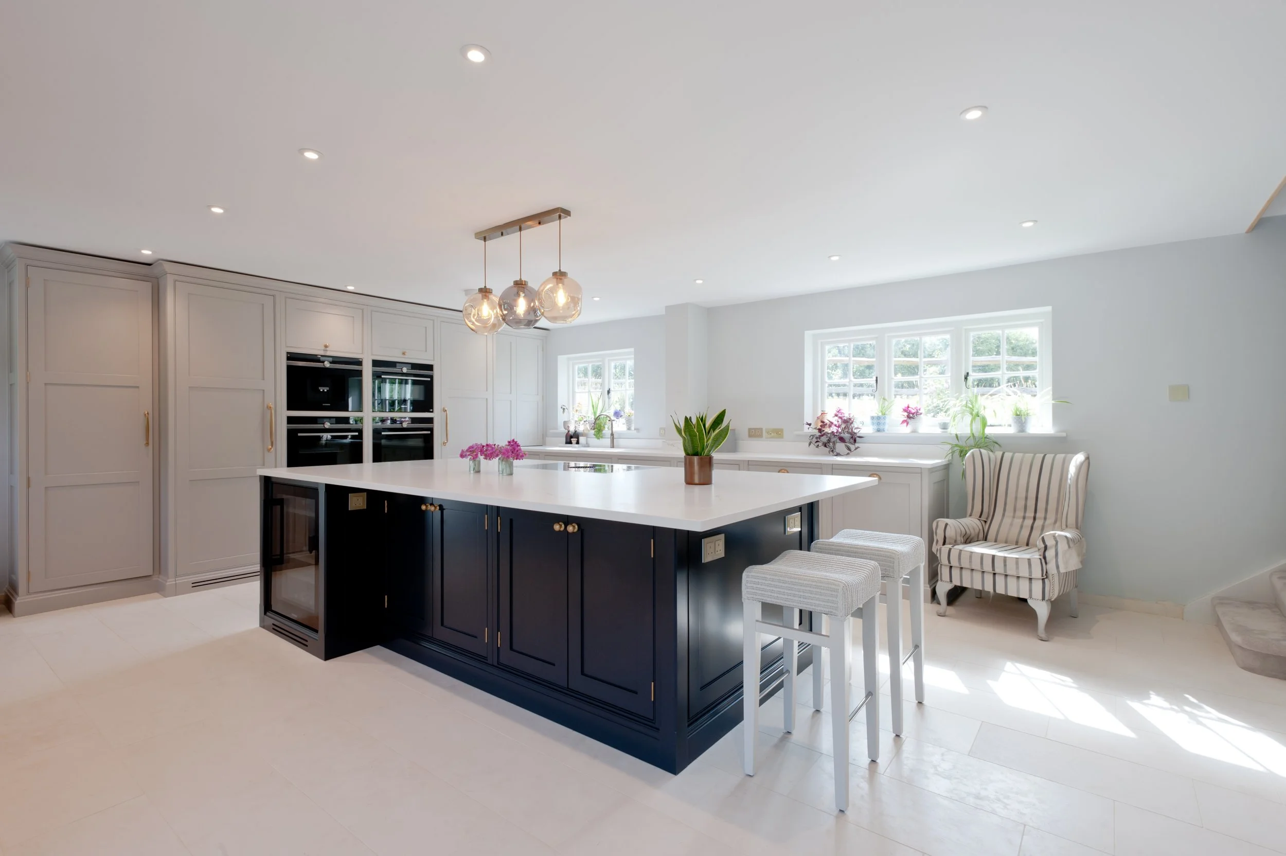 Modern kitchen with a black island, white countertops, built-in oven, and gray cabinets. Well-lit with ceiling spotlights, a hanging light fixture with bulbs, and natural light from two windows. Decor includes potted plants and striped armchair.