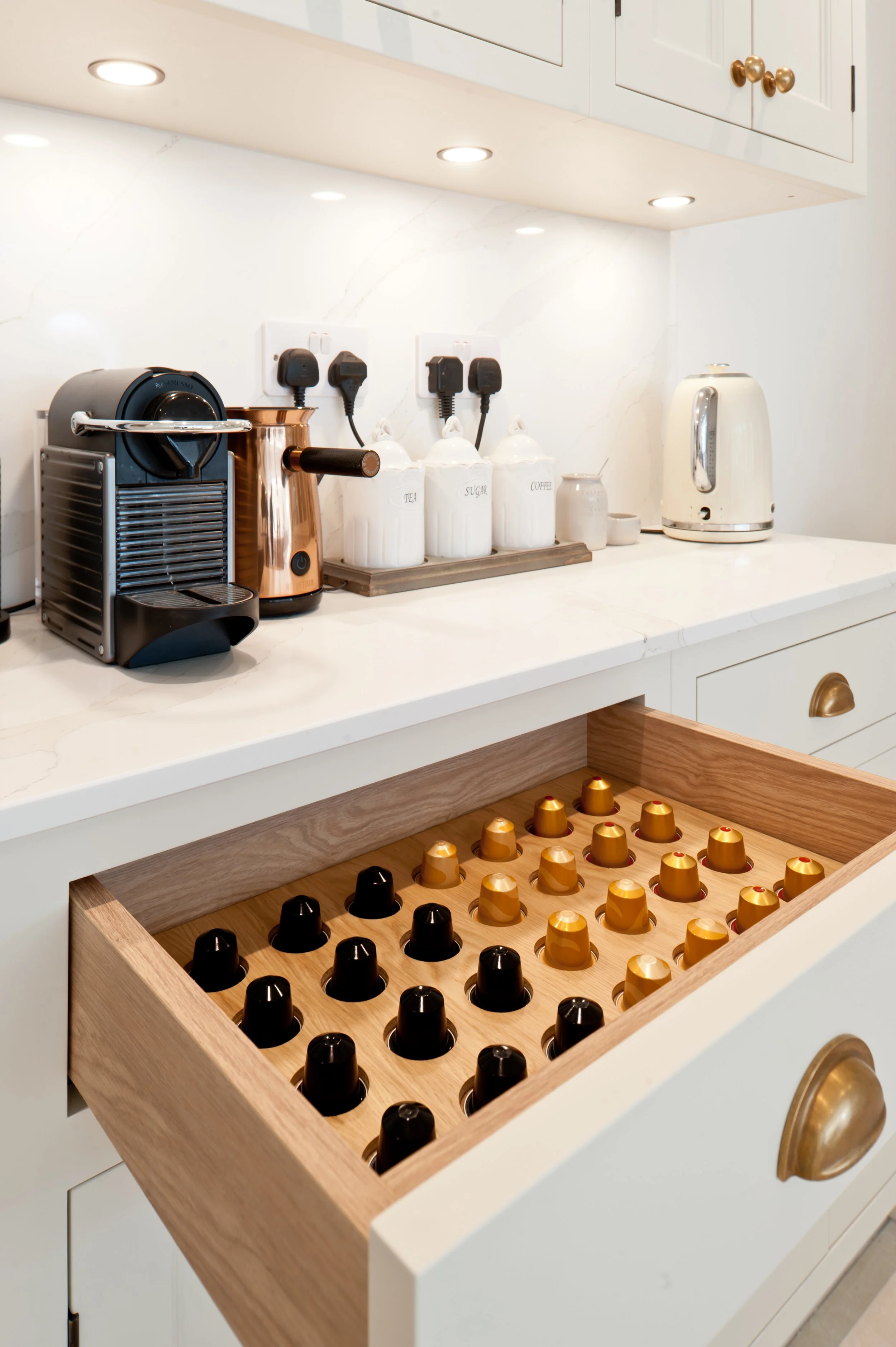 Open drawer with black and gold coffee capsules inside, part of kitchen countertop with coffee and tea canisters, electric kettle, and coffee machine.