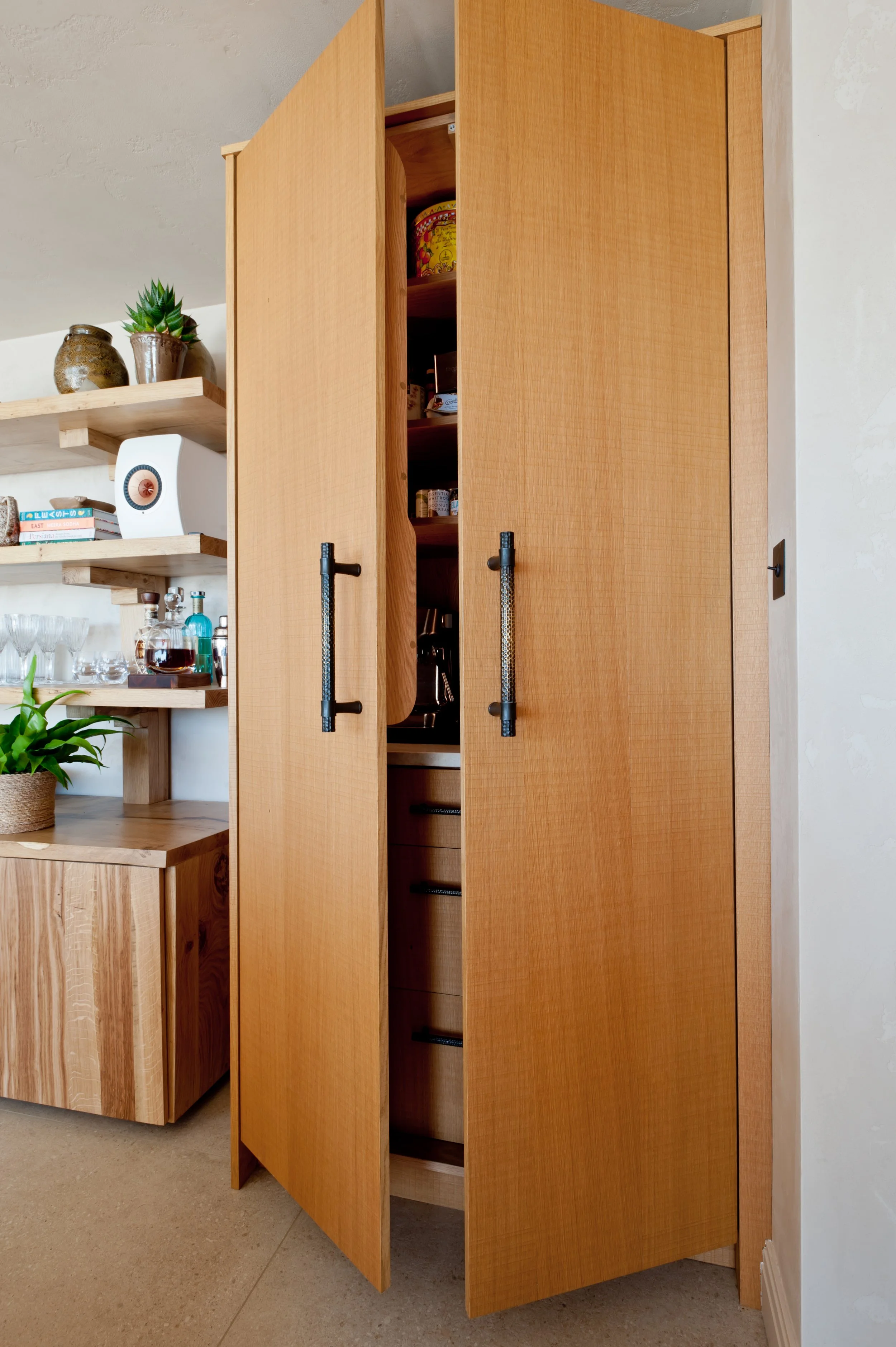 A wooden kitchen cabinet with two doors partially open, revealing shelves inside. To the left, there are wooden shelves with potted plants, books, and glassware.