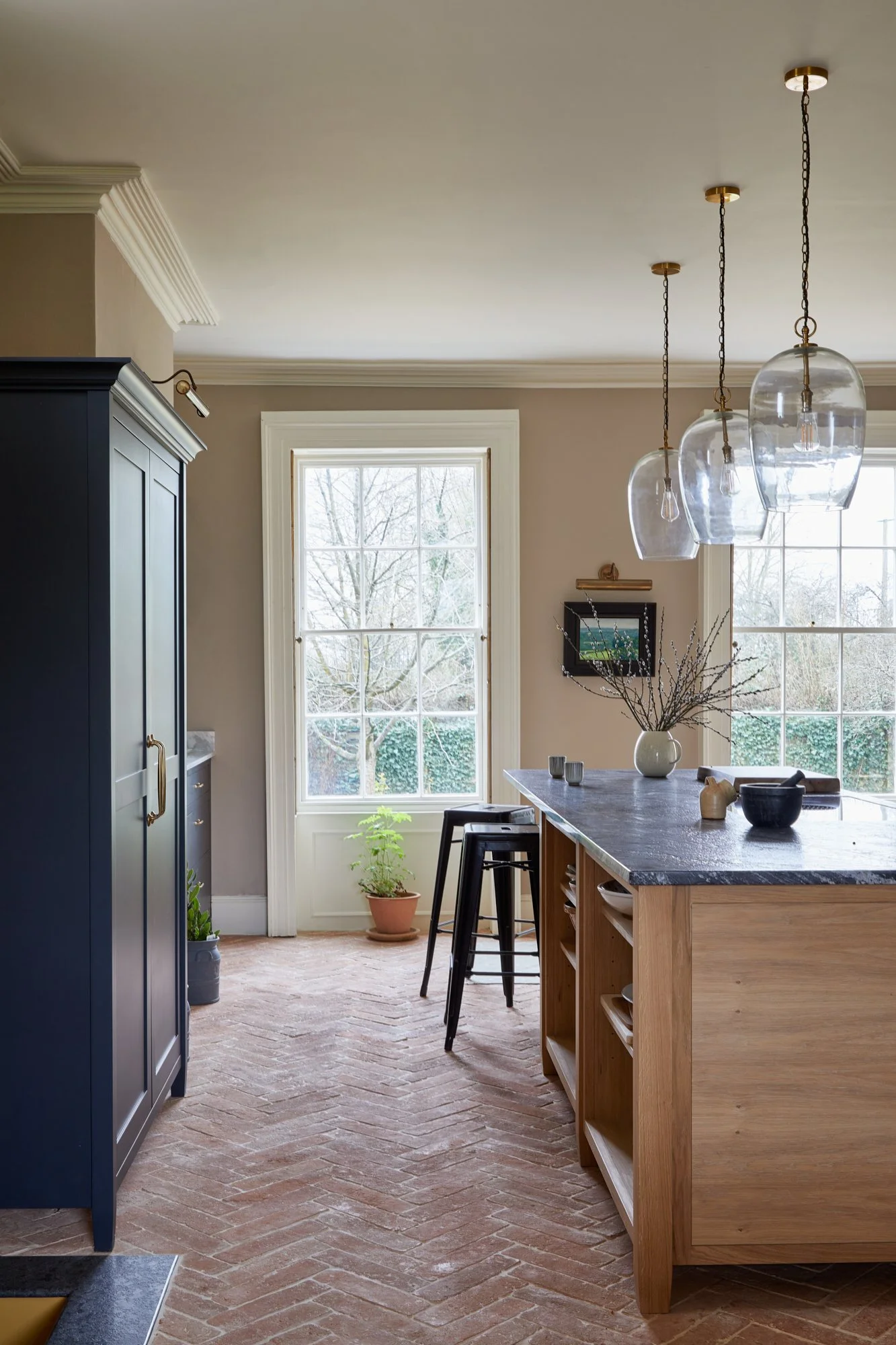 A bright kitchen with large windows, a wooden kitchen island with dark gray countertops, black bar stools, a navy cabinet, and decorative items including a vase with branches and small bowls.