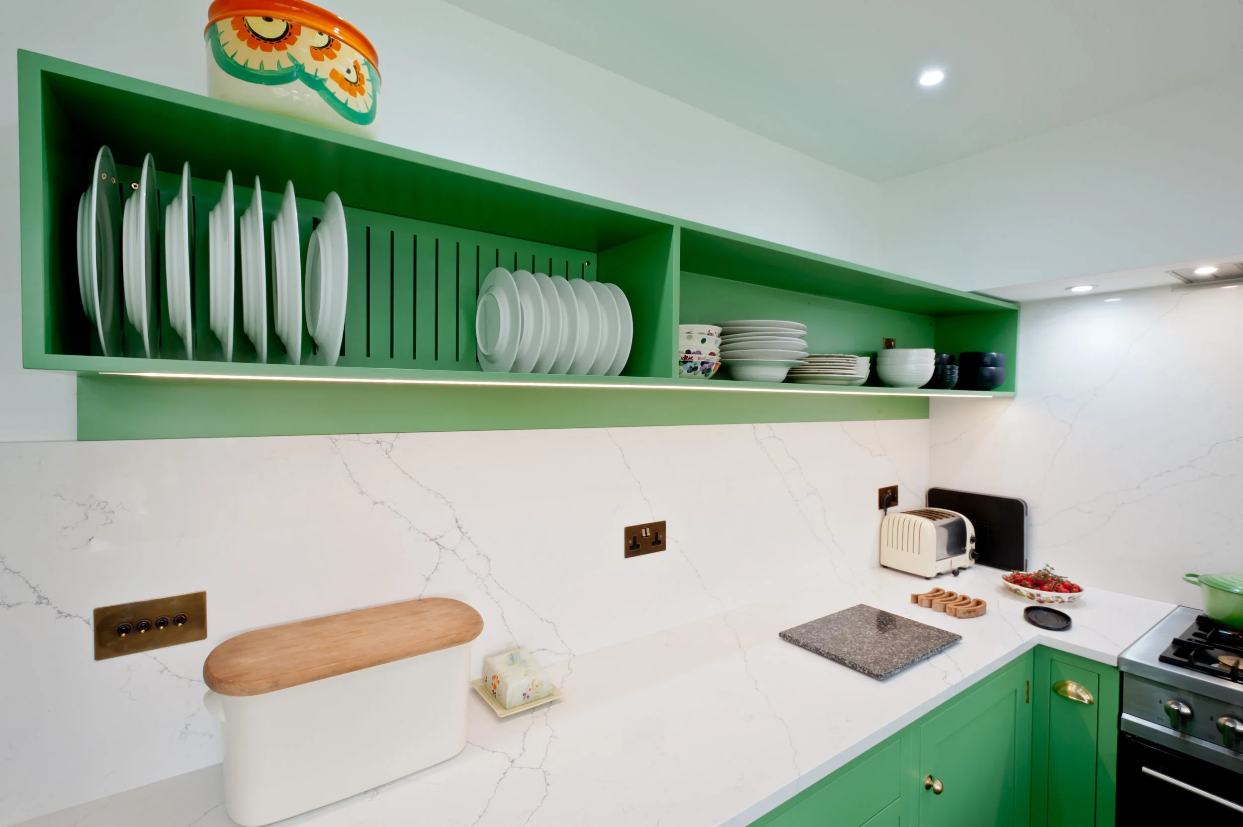 Green kitchen shelf with white dishes, bowls, and black cups, white marble countertop, toaster, and stove, with a white wall and marble backsplash.