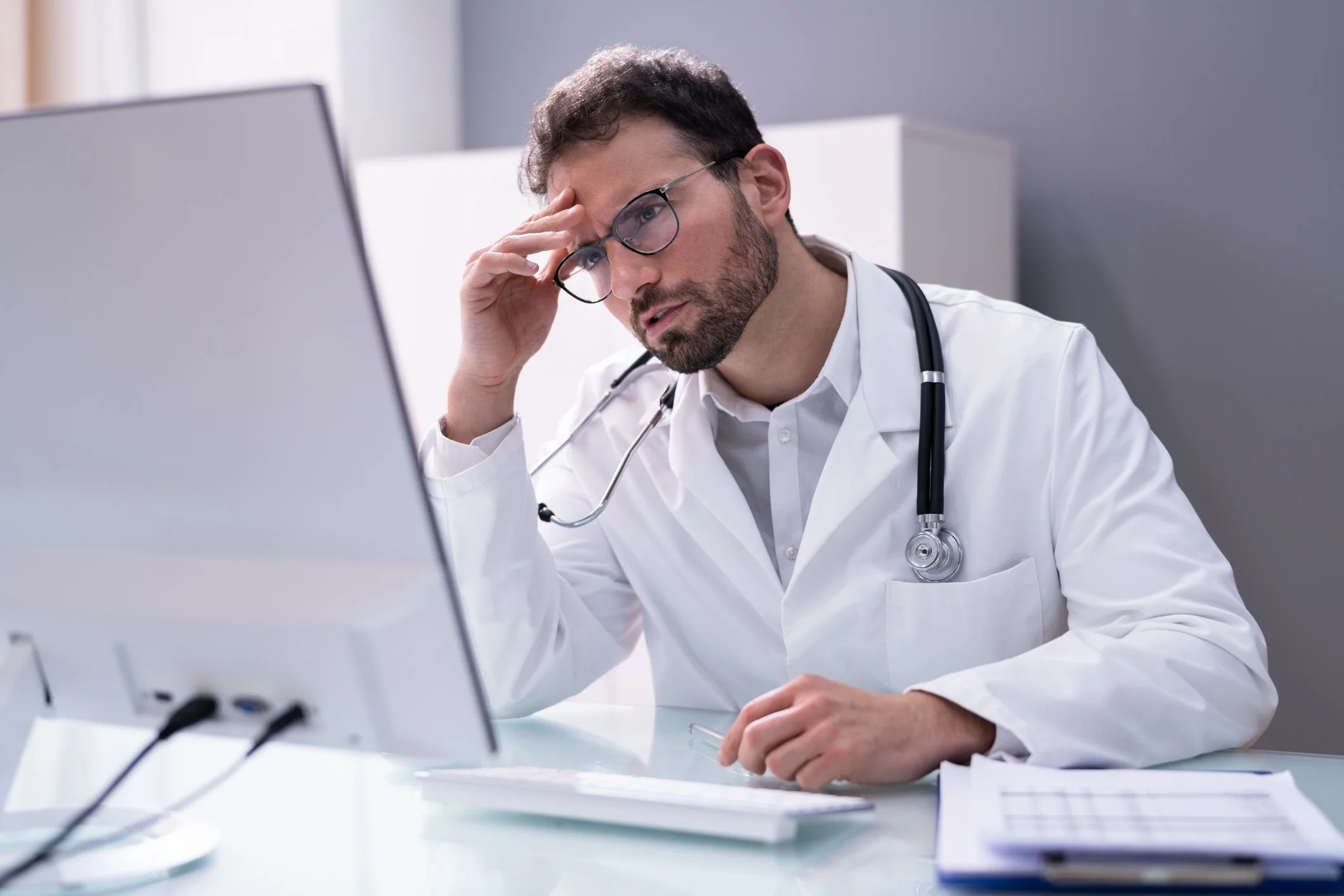 A male doctor wearing glasses and a white lab coat, sitting at a desk looking frustrated while holding his hand to his forehead, with a stethoscope around his neck, in a medical office or clinic.