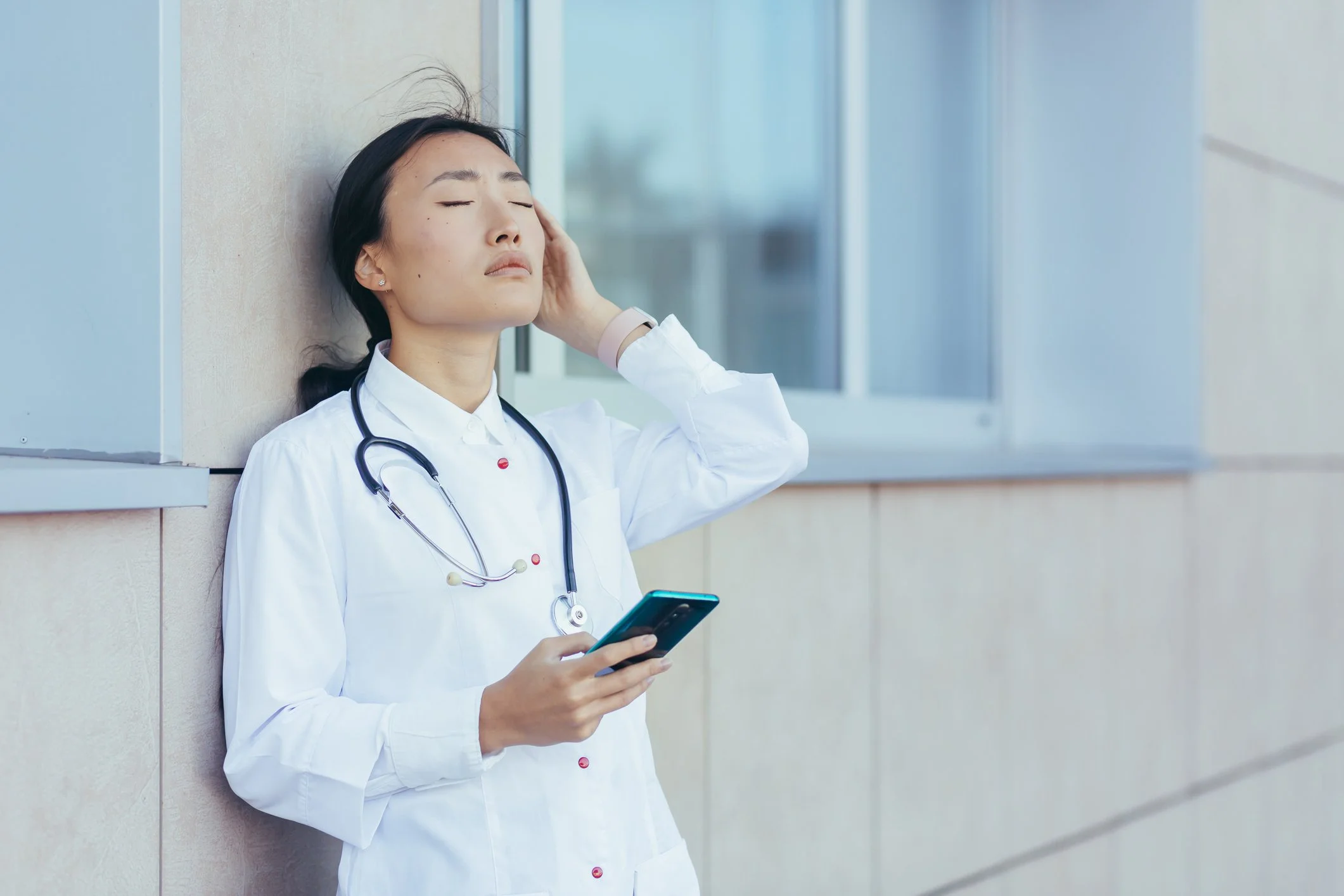 A female healthcare professional in a white coat and stethoscope looking at her phone while leaning against a wall, with her eyes closed and her hand resting on her head.