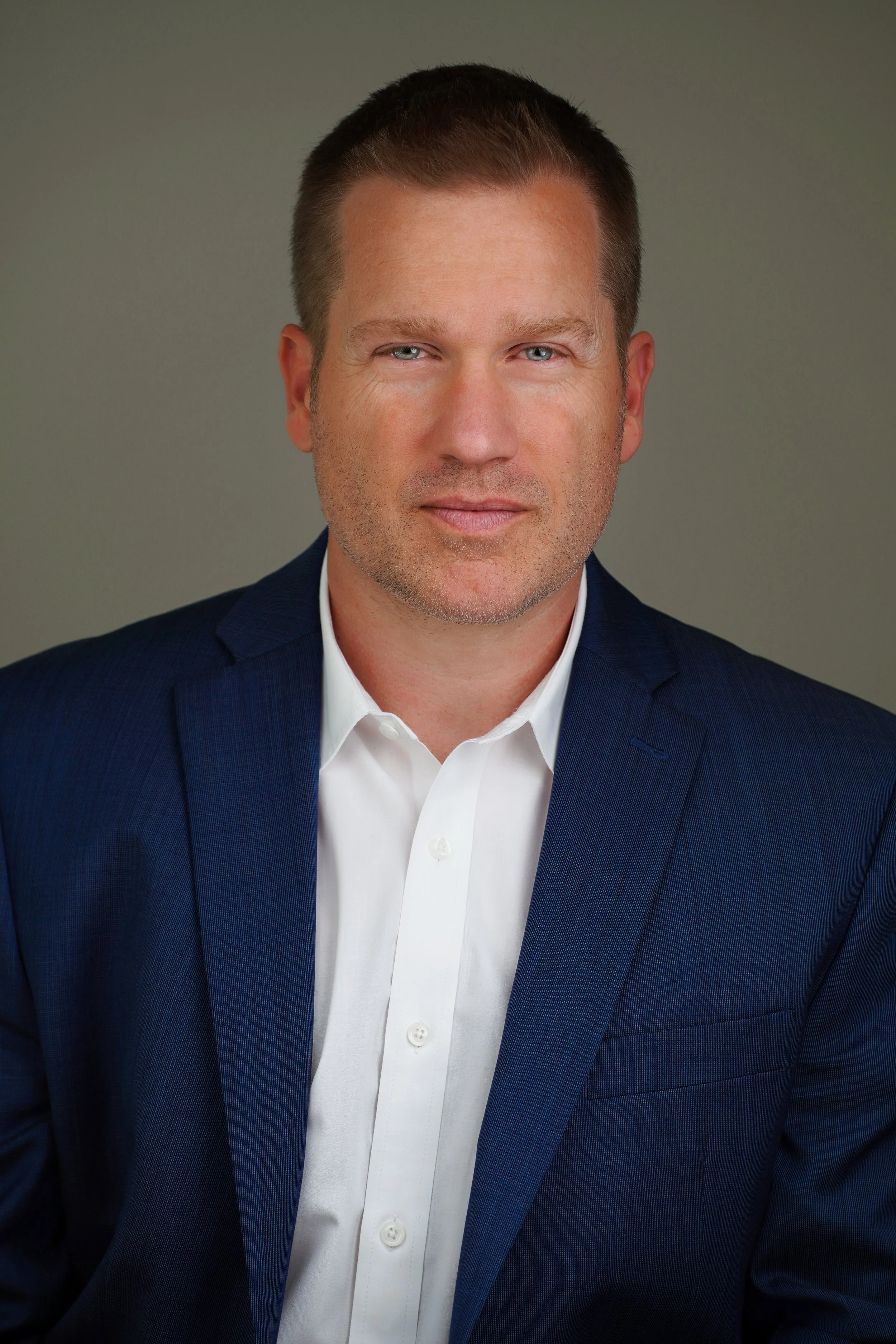 Headshot of a man with short brown hair, blue eyes, wearing a white dress shirt and a dark blue suit jacket, against a gray background.