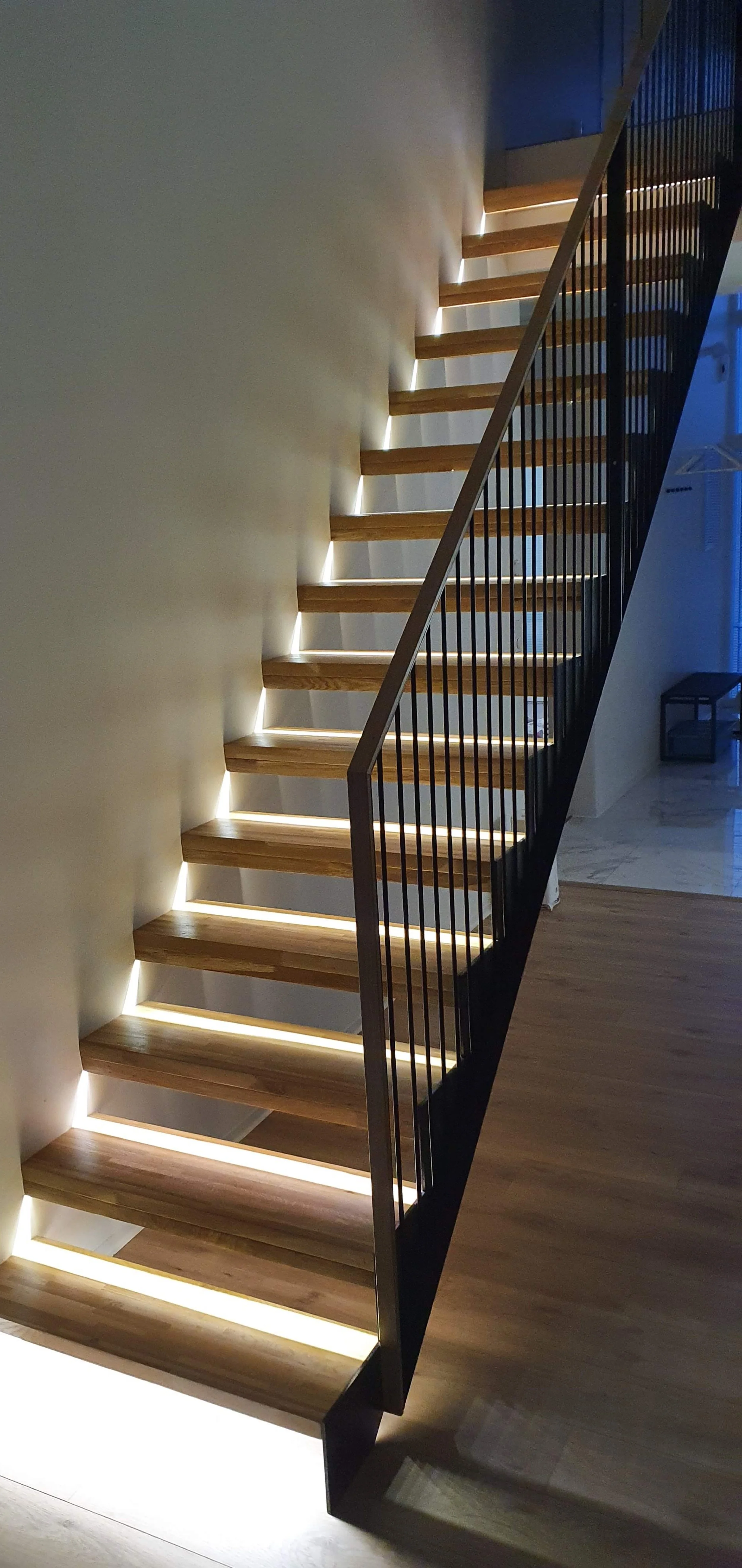 Modern staircase with wooden steps and LED lighting underneath each step, black metal railing on the right side, situated in a contemporary interior space.
