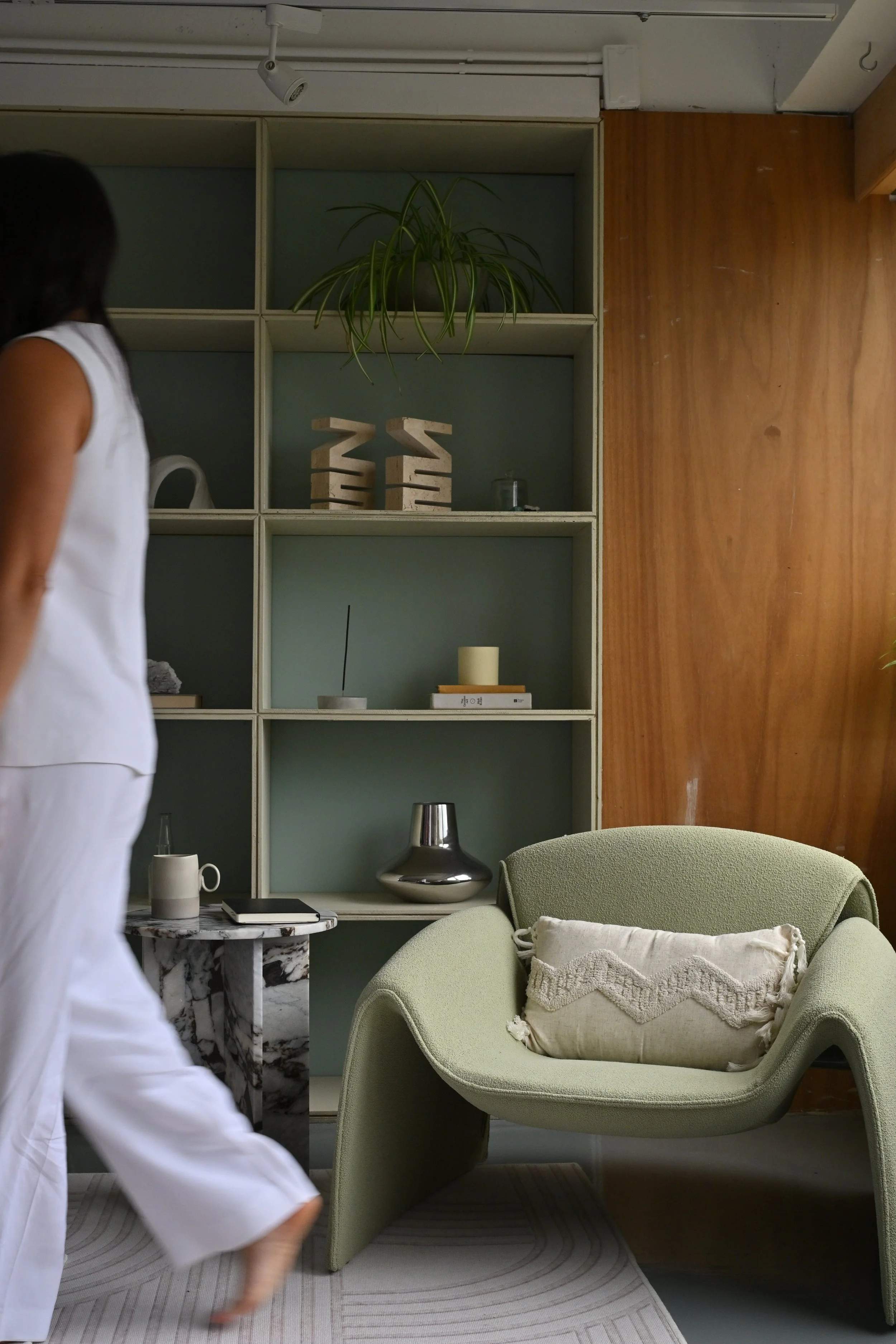 Interior view of a cozy living room with a green modern armchair with a beige cushion, a white bookshelf with decorative items, a marble side table with a cup and a notebook, a small plant on the top shelf, and a wooden wall.