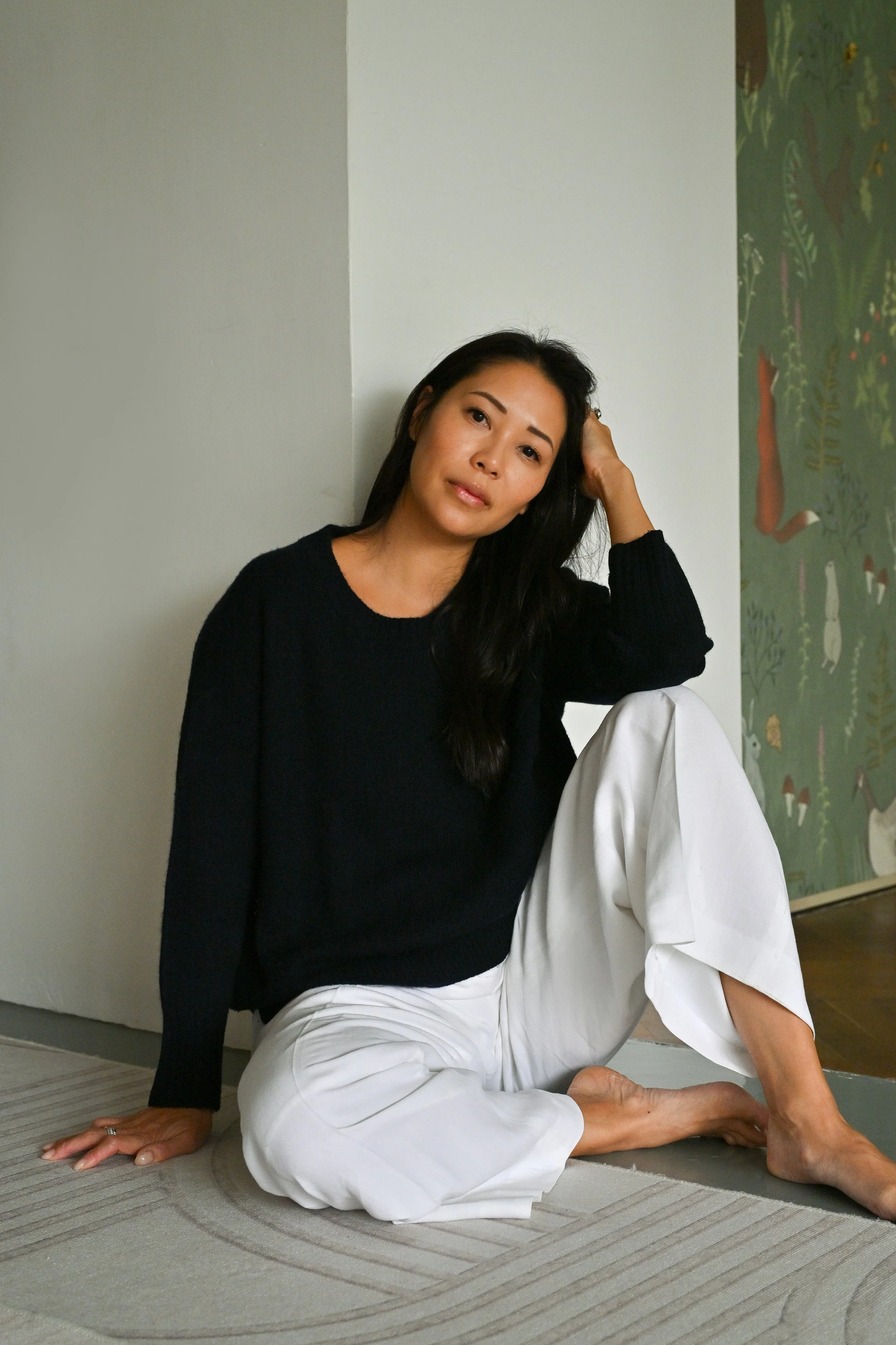 Woman in black sweater and white pants performing a yoga pose on a mat inside a room, with a painted wall featuring nature-themed artwork in the background.
