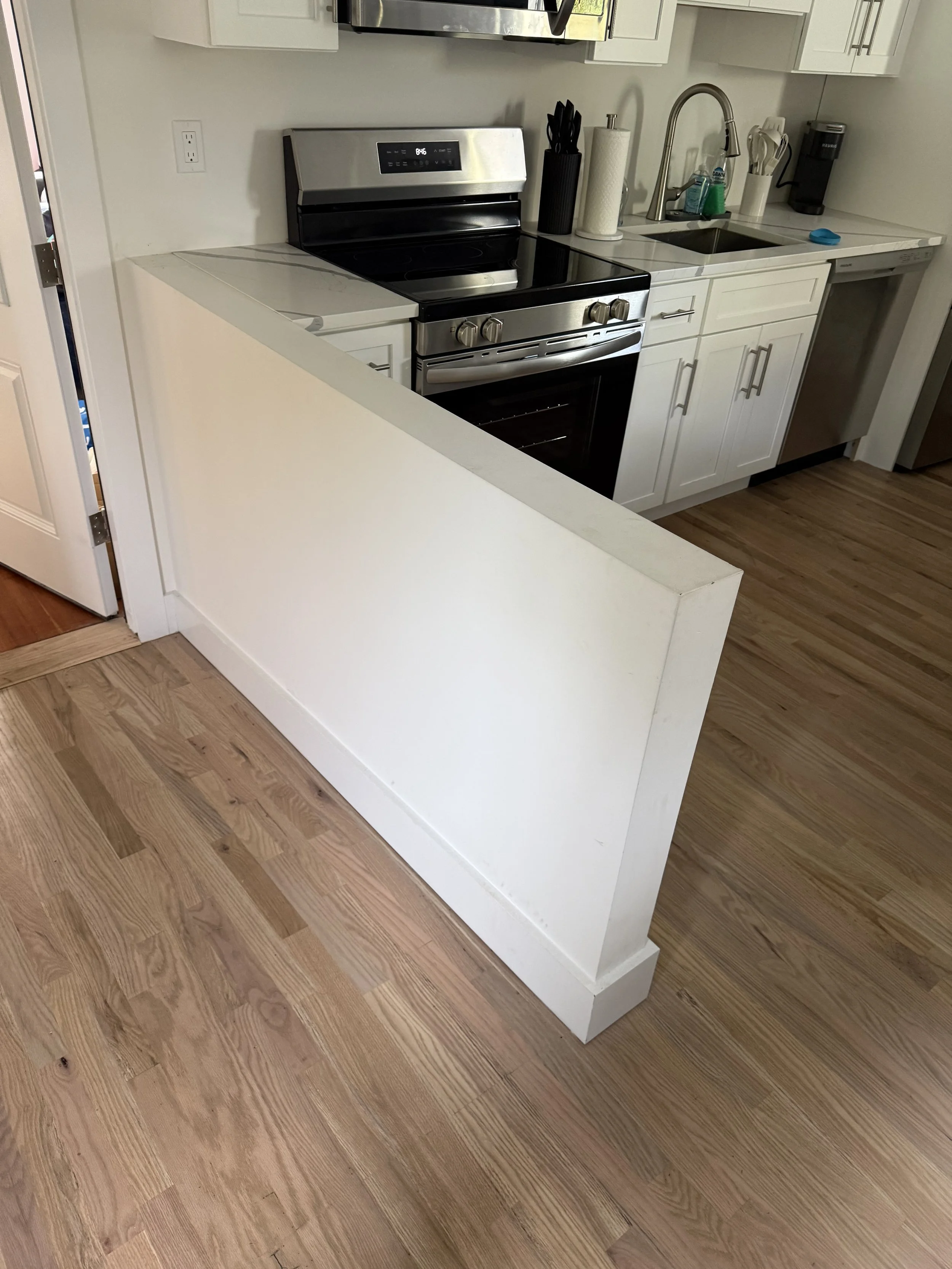 A modern kitchen with white cabinets, a stainless steel stove and oven, a white countertop, and a sink. Items on the counter include a black utensil holder, paper towels, a dish soap bottle, a dish scrubber, and a black coffee maker. Hardwood floorin