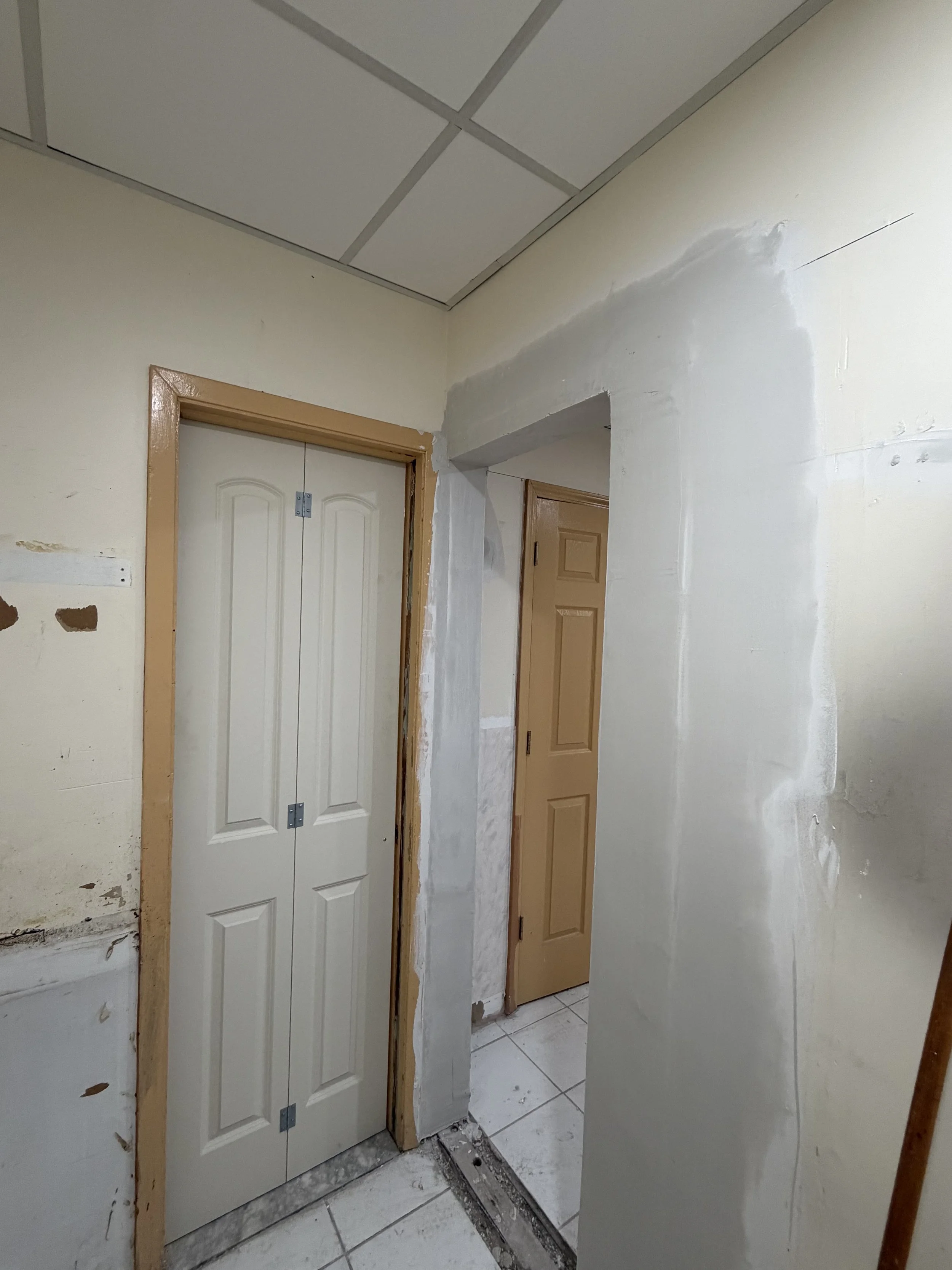 Interior hallway with unfinished walls, two closed doors, tile floor, ceiling tiles, and construction work in progress.