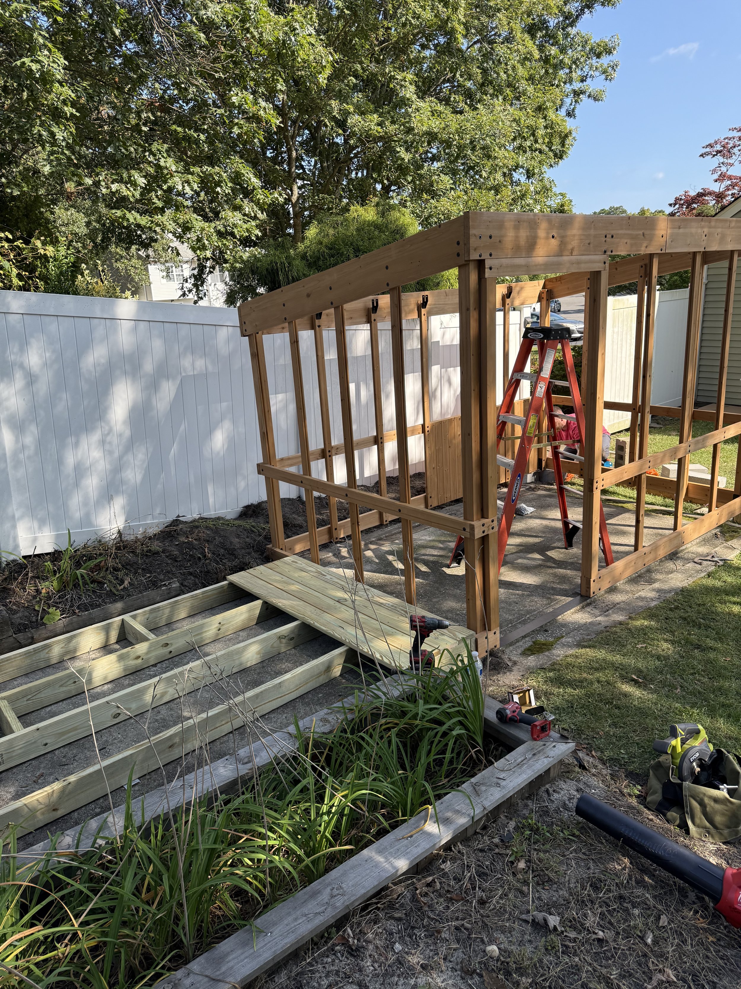 A backyard in the process of construction with a wooden structure frame, a red ladder, tools, and garden beds.