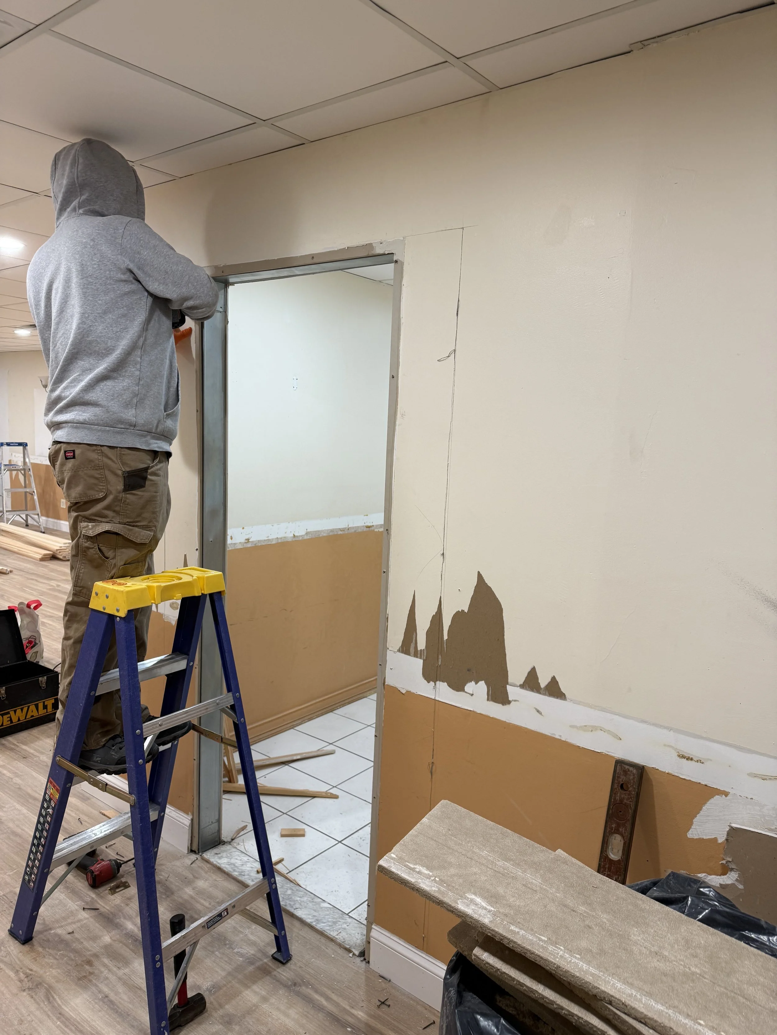 A person in a gray hoodie and tan cargo pants standing on a ladder, working on a wall with a doorway under renovation. The wall has patches of brown paint and exposed drywall with a level tool leaning against it. Construction tools and debris are vis