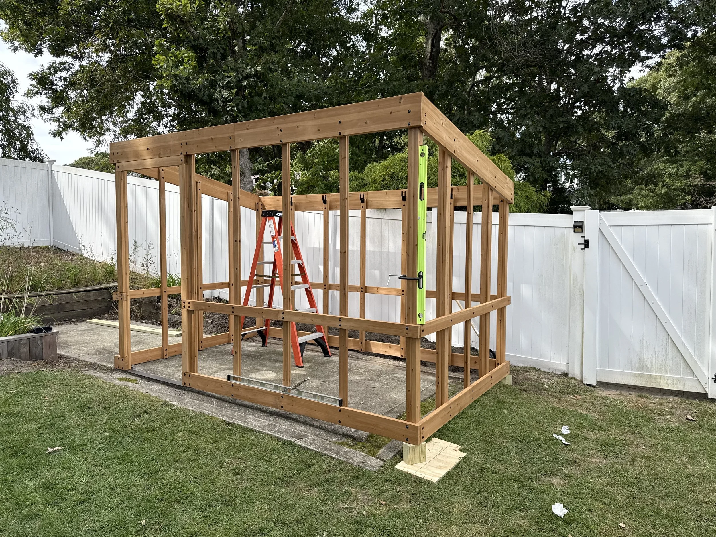 Wooden frame structure under construction in a backyard, with a red step ladder, surrounded by a white fence and with trees in the background.