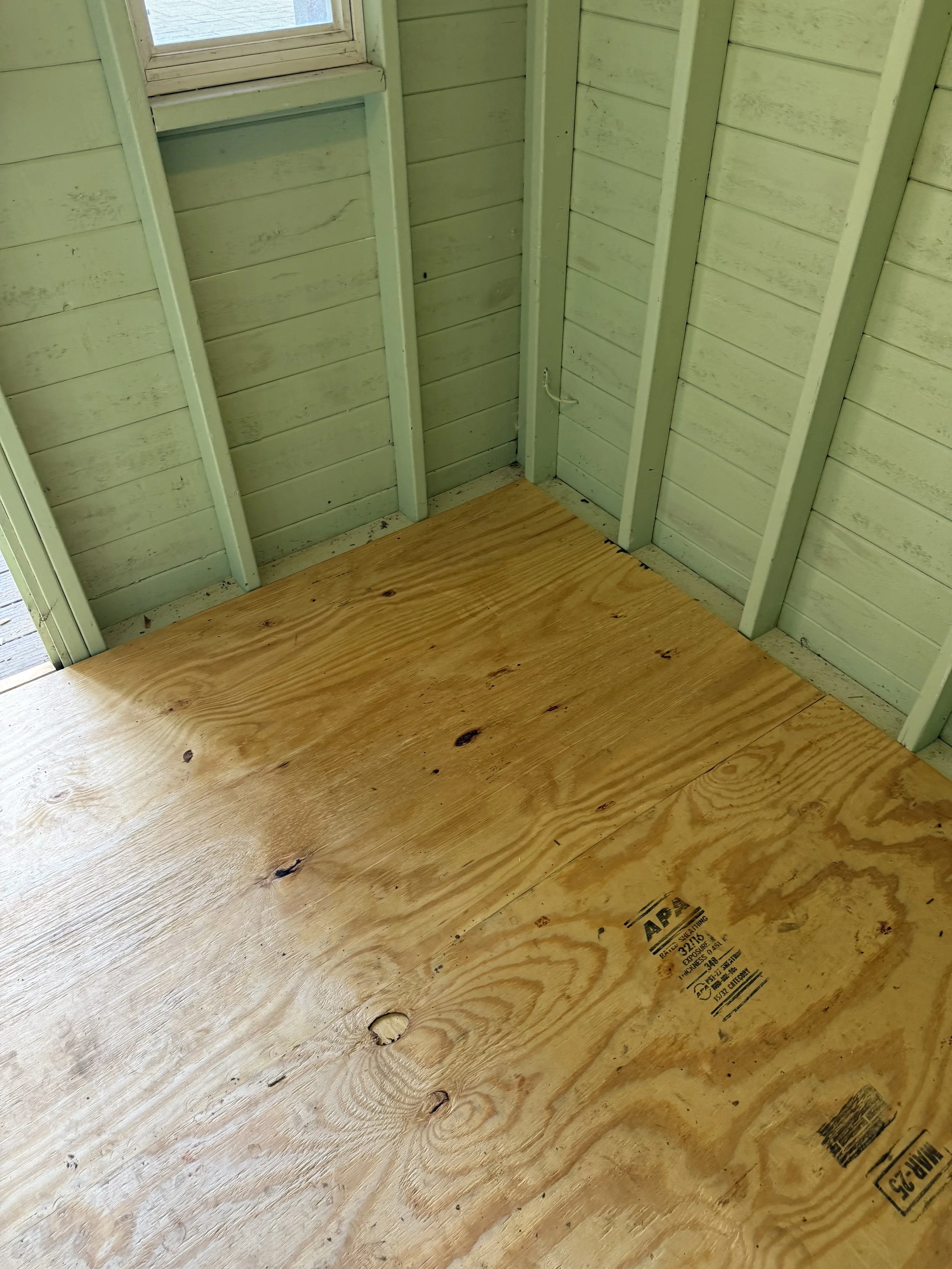 Interior of a small shed or room with light green painted wooden walls and a partly visible window. The floor is made of unfinished plywood panels.