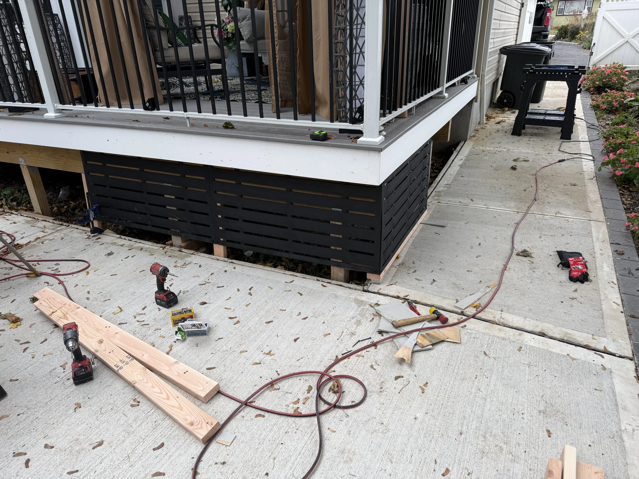 Construction site on a house's porch with tools, wood pieces, and a saw. The porch has a black railing and a garden with pink flowers nearby.