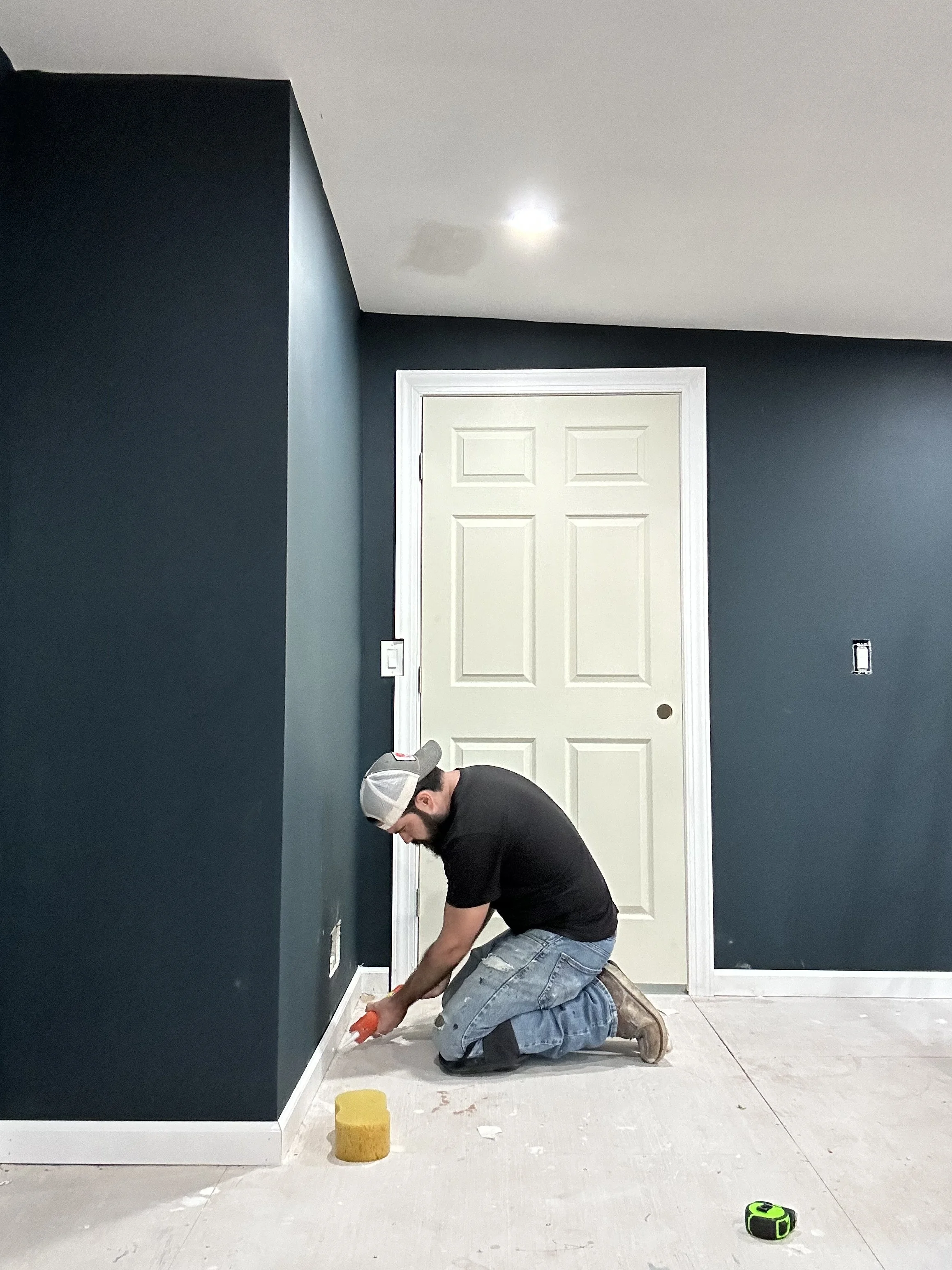 A man kneeling on the floor, applying baseboard molding to a dark teal wall with tools nearby, in a room under renovation.