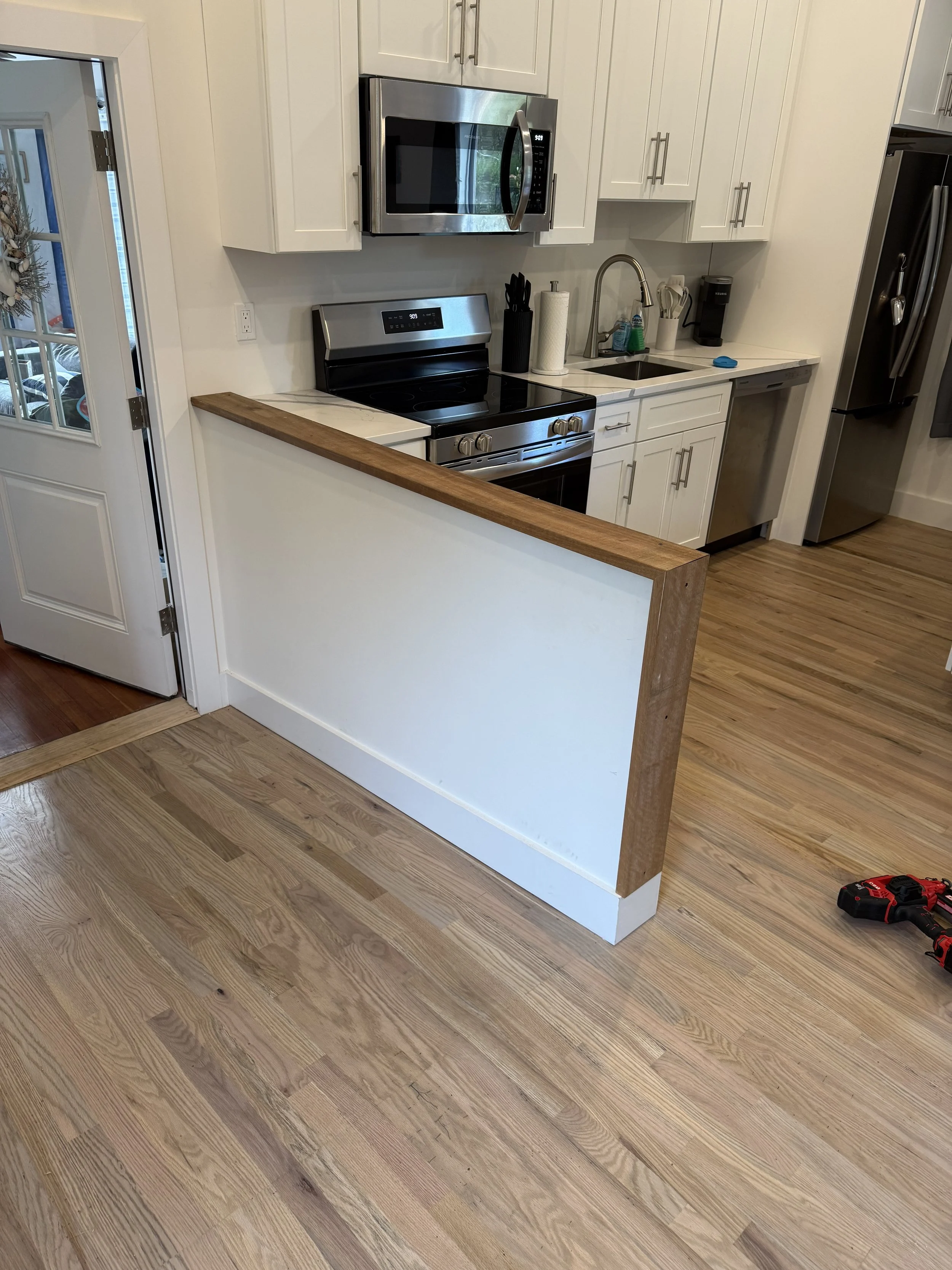 A modern kitchen with white cabinets, stainless steel appliances, including a microwave and oven, and a wooden countertop. There is a small breakfast bar or partial wall, with hardwood flooring and some tools on the floor.
