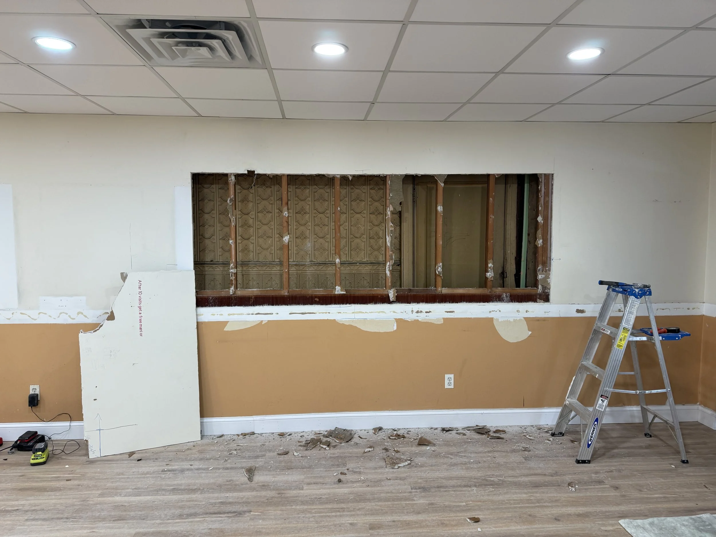 Room undergoing renovations with a hole in the wall exposing the interior framing. Construction tools and debris are on the floor, and a silver ladder stands to the right.