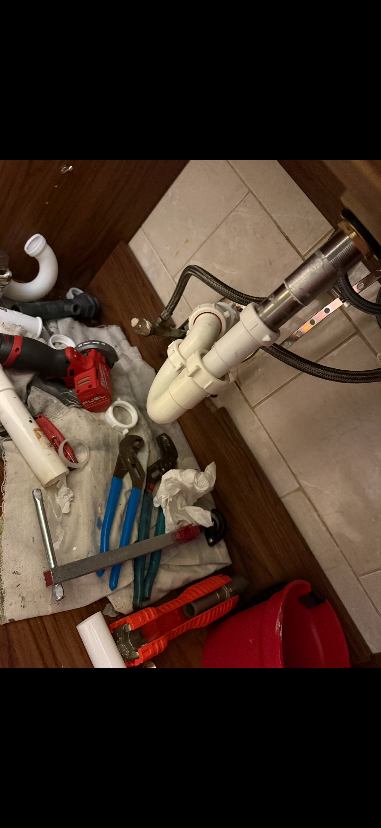 Under the kitchen sink, plumbing repair tools and parts including white PVC pipes, a pipe wrench, a hacksaw, a cordless drill, adjustable pliers, and a red toolbox are scattered on a cloth on the wooden floor.