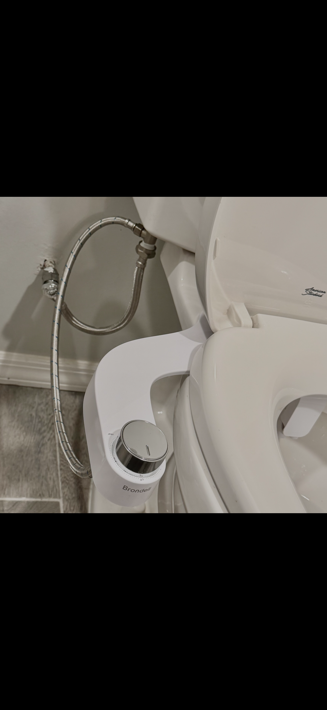 Toilet with a Brondell bidet attachment and metallic water supply hoses connected to the wall.