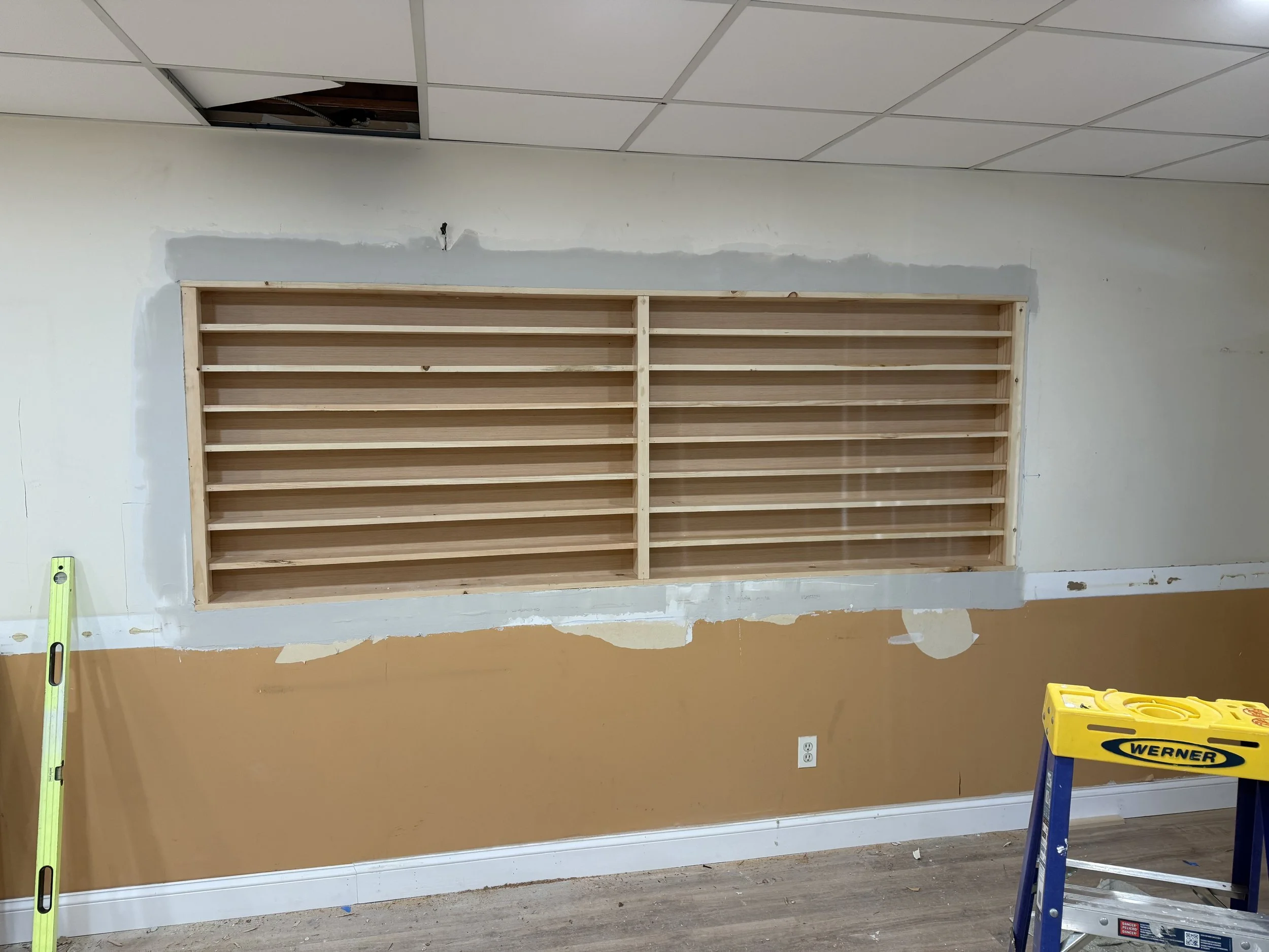 A wall with a rectangular framed section containing wooden shelves and a missing ceiling tile above, with construction tools and materials nearby.