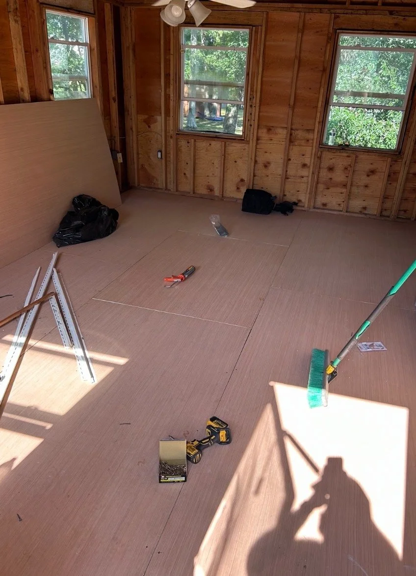 Interior of a room under construction with unfinished wooden walls, two windows, and construction tools and materials on the floor.