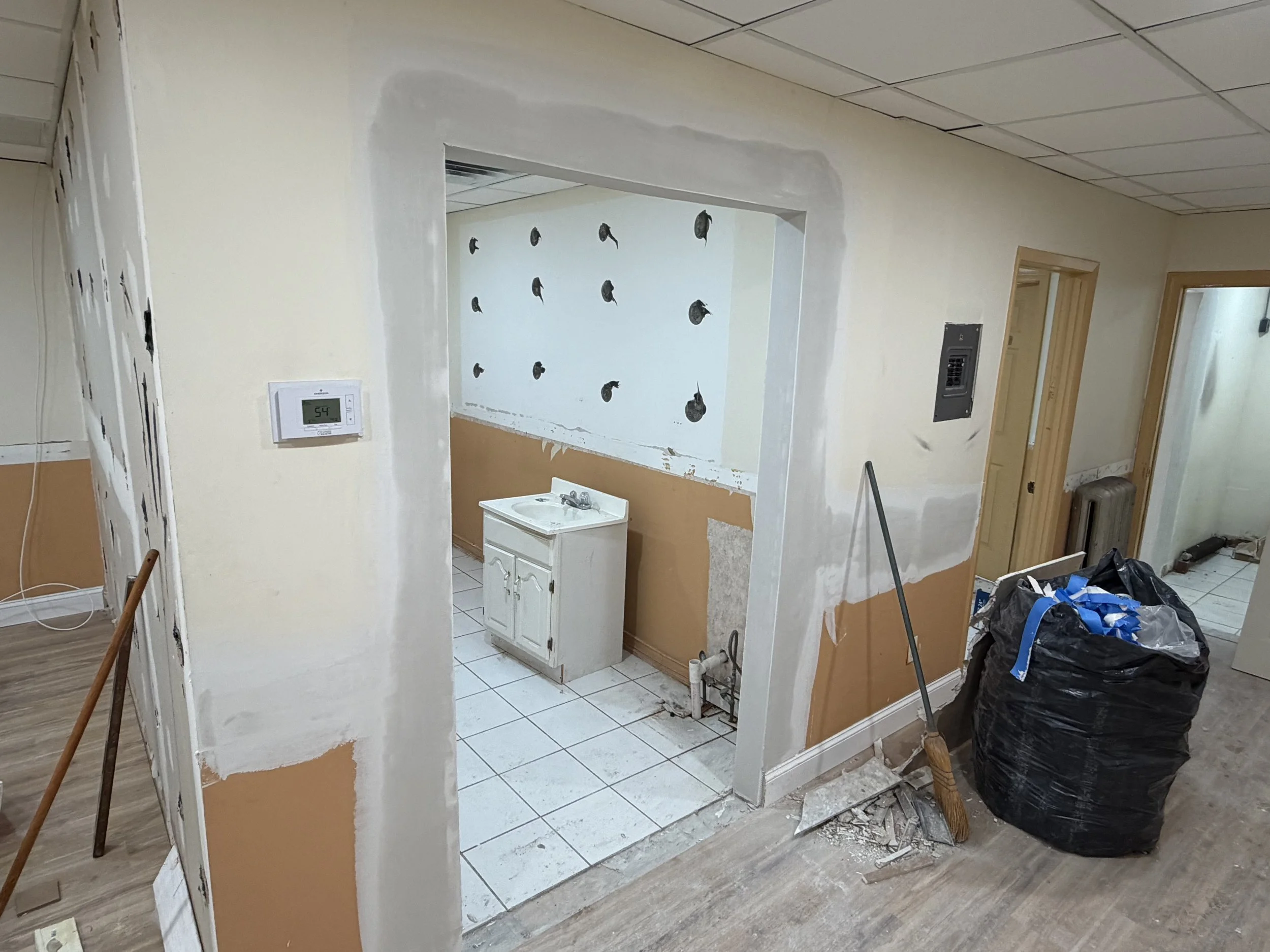 Room under renovation with a small white sink cabinet, unfinished walls, and construction materials including a large black trash bag, broom, and dust debris.