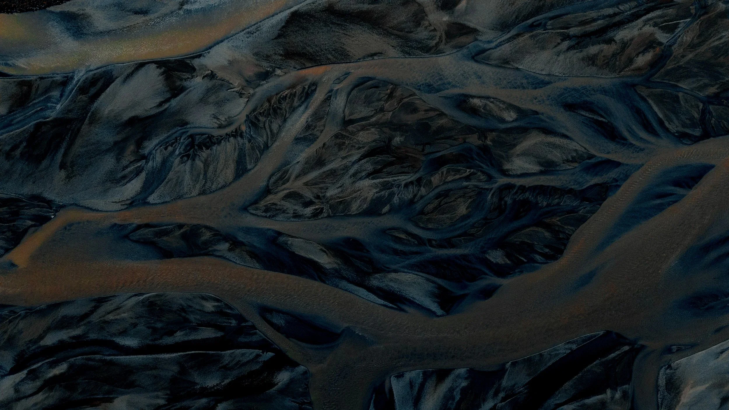 An aerial view of dark, winding river channels surrounded by barren, rocky terrain at sunset.