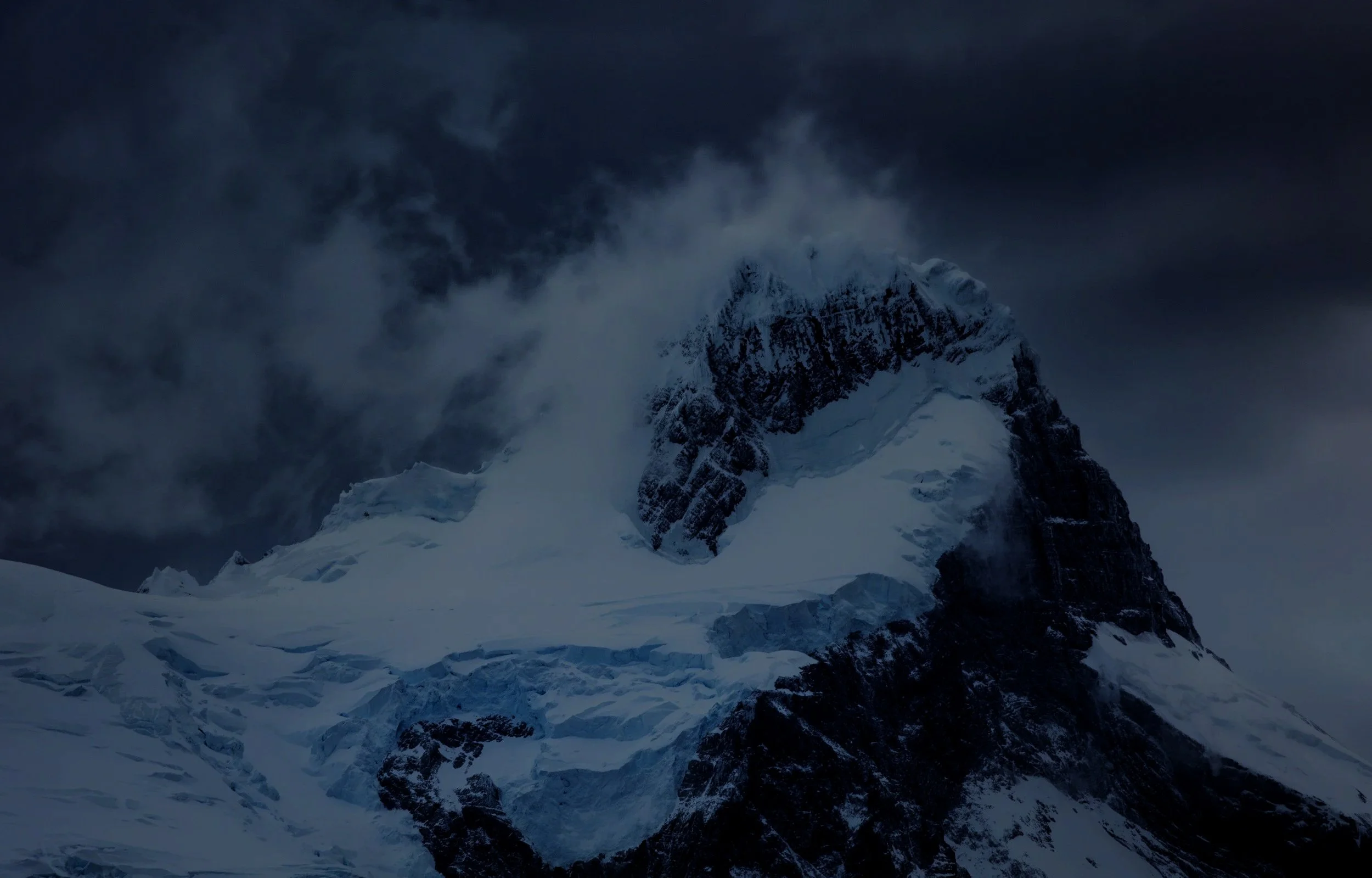Snow-covered mountain peak with clouds and dark sky in the background.