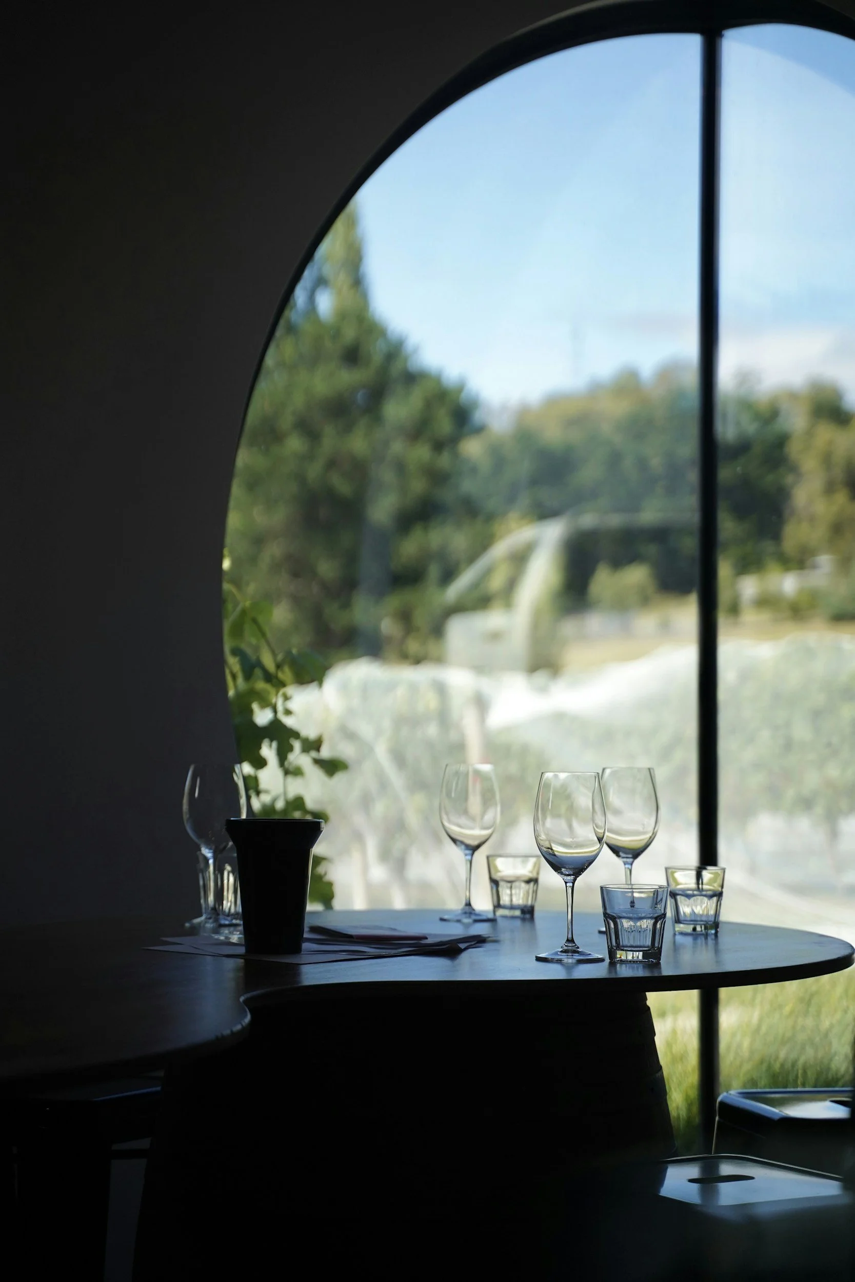 Empty wine glasses and water glasses on a table near a large window with a view of trees and a blue sky.