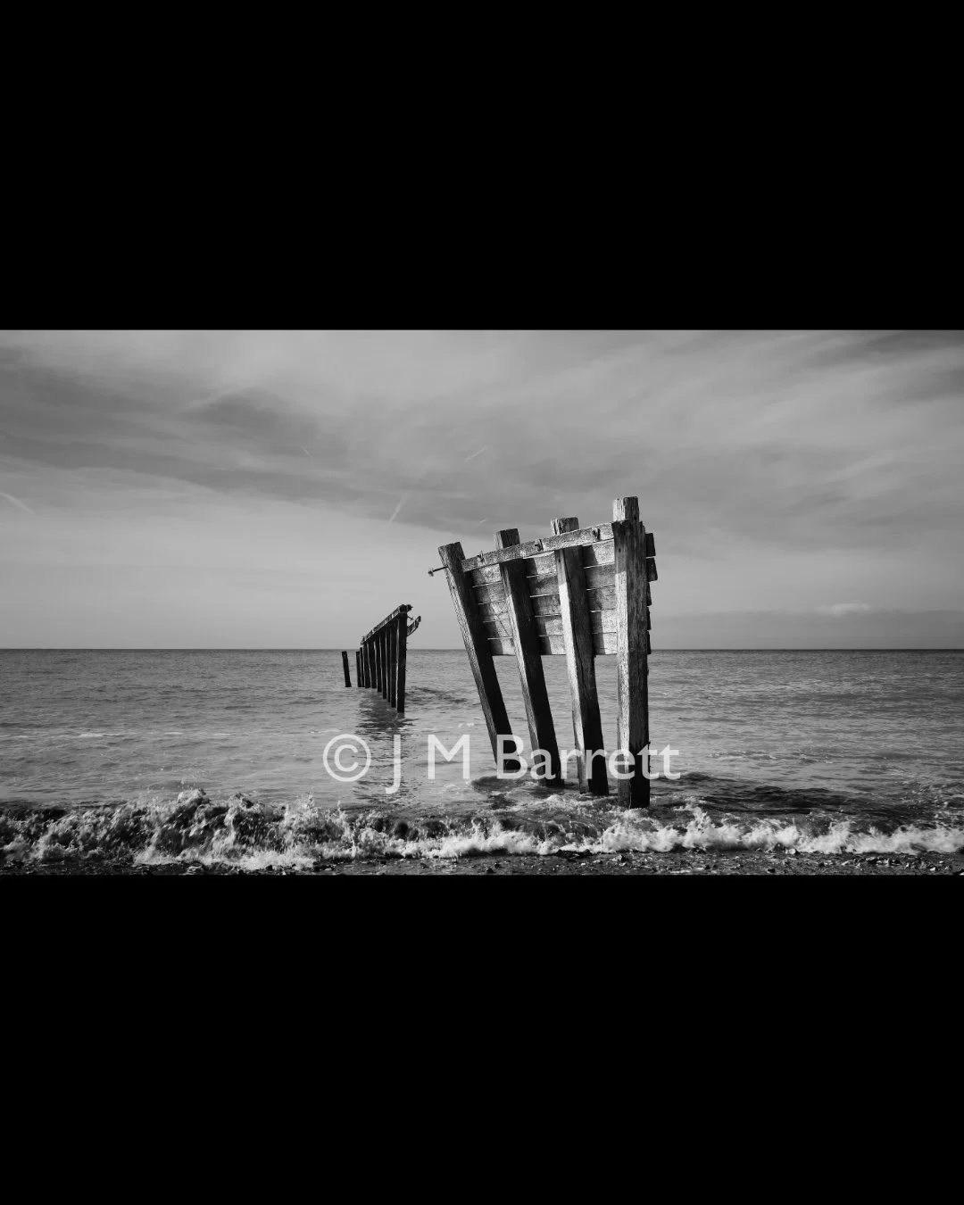 Black and white photograph of a partially submerged, weathered shipwreck on the beach with the ocean in the background and a cloudy sky.