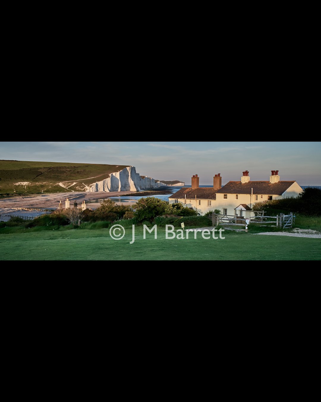 Cliffs on the coast with white chalk formations, a row of houses with chimneys, and green vegetation at sunset.