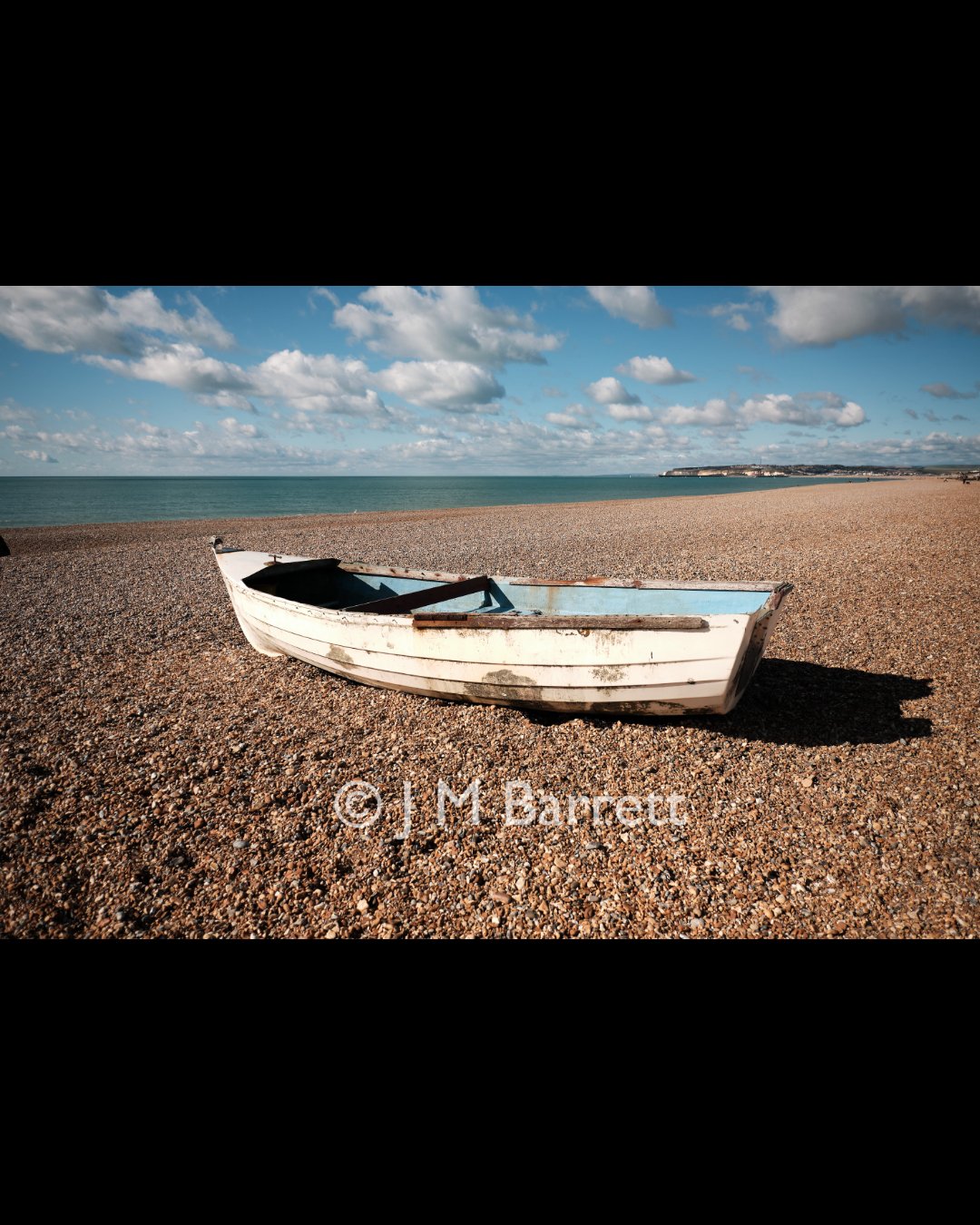 Old white boat resting on a pebble beach with the ocean and a cloudy sky in the background.