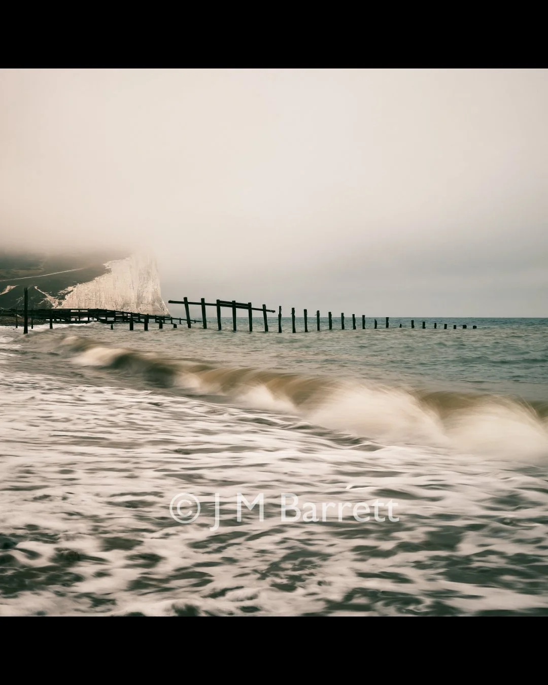 Photo of a beach with broken wooden groyne and white chalk cliffs in the background, cloudy sky, ocean waves in the foreground.
