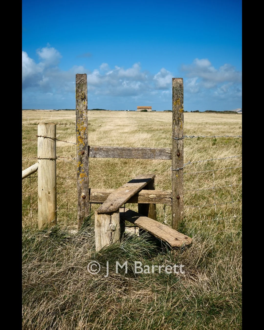 A weathered wooden step ladder leading up to a fence in a grassy field, with a small building in the distance under a partly cloudy blue sky.