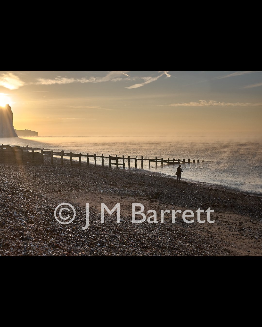 Sunset over a pebble beach with a person fishing near the water, and a wooden groyne extending into the sea. Cliffs are visible in the distance.