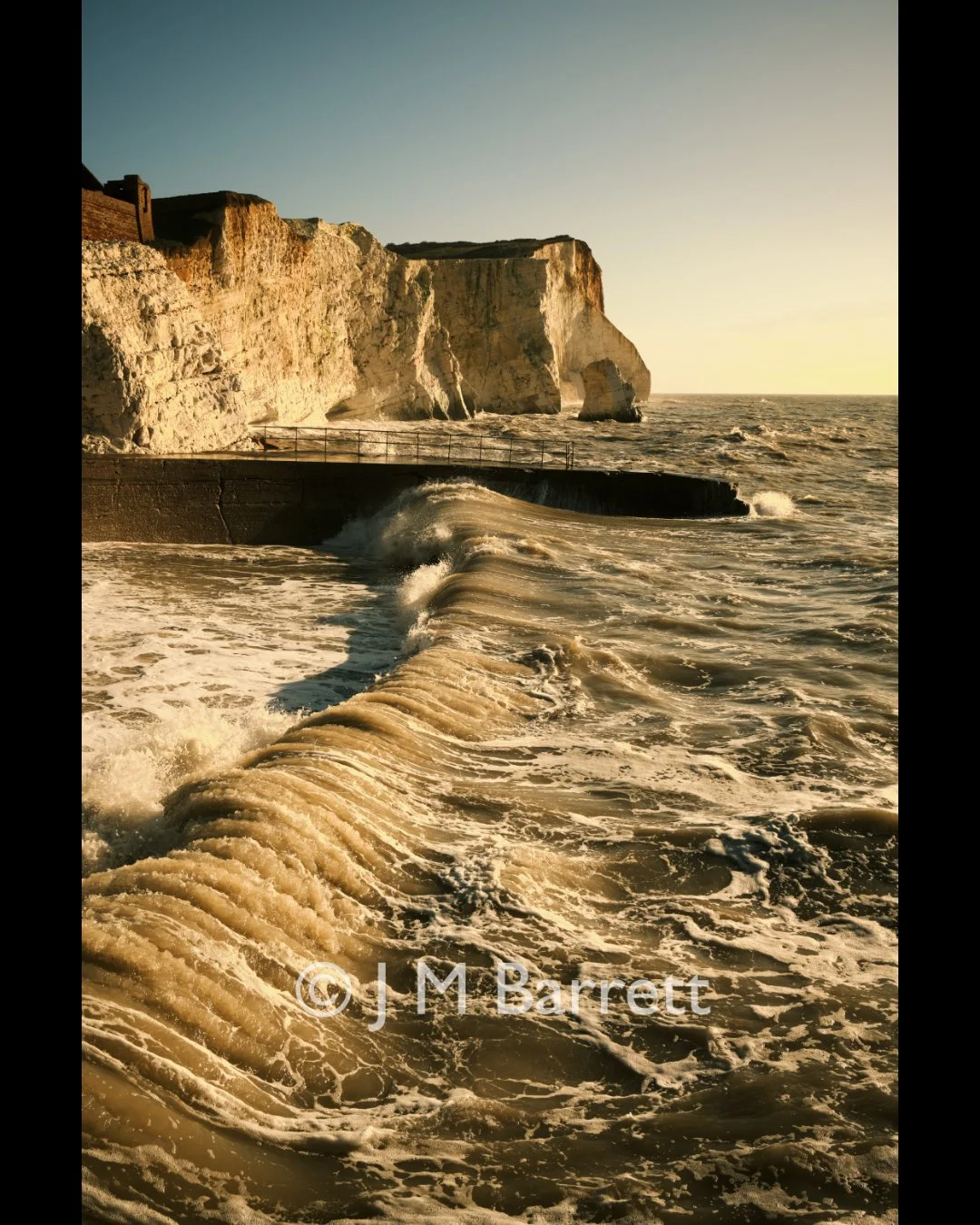 Cliffs overlooking the ocean at sunset, with waves crashing against a concrete barrier.