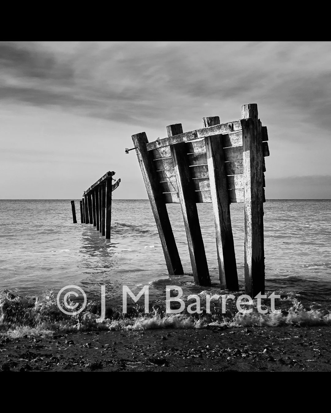 Black and white photo of a decaying wooden boat wreck on a beach with the ocean in the background.