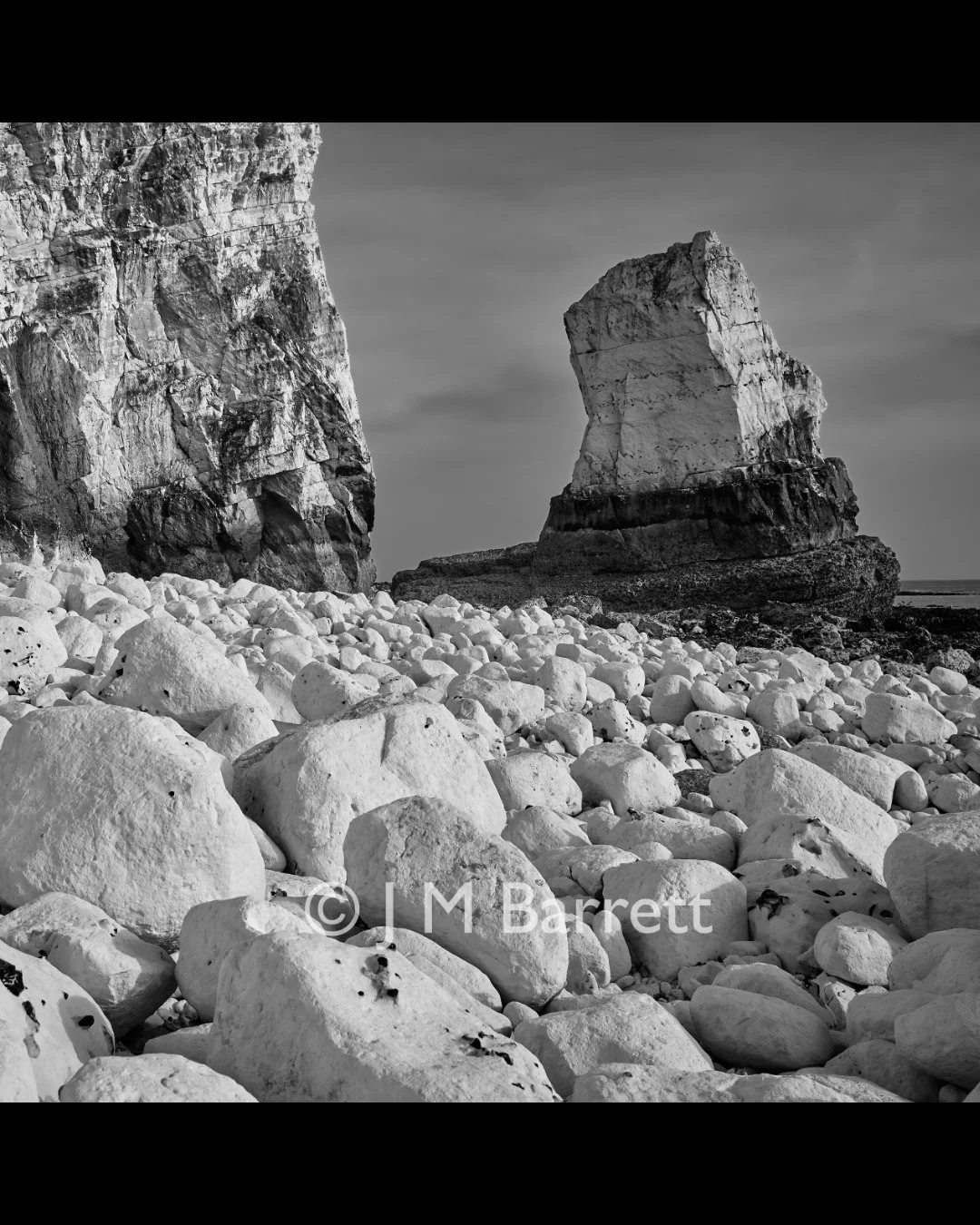 Black and white photo of rocky beach with large white stones and towering chalk cliffs in the background.