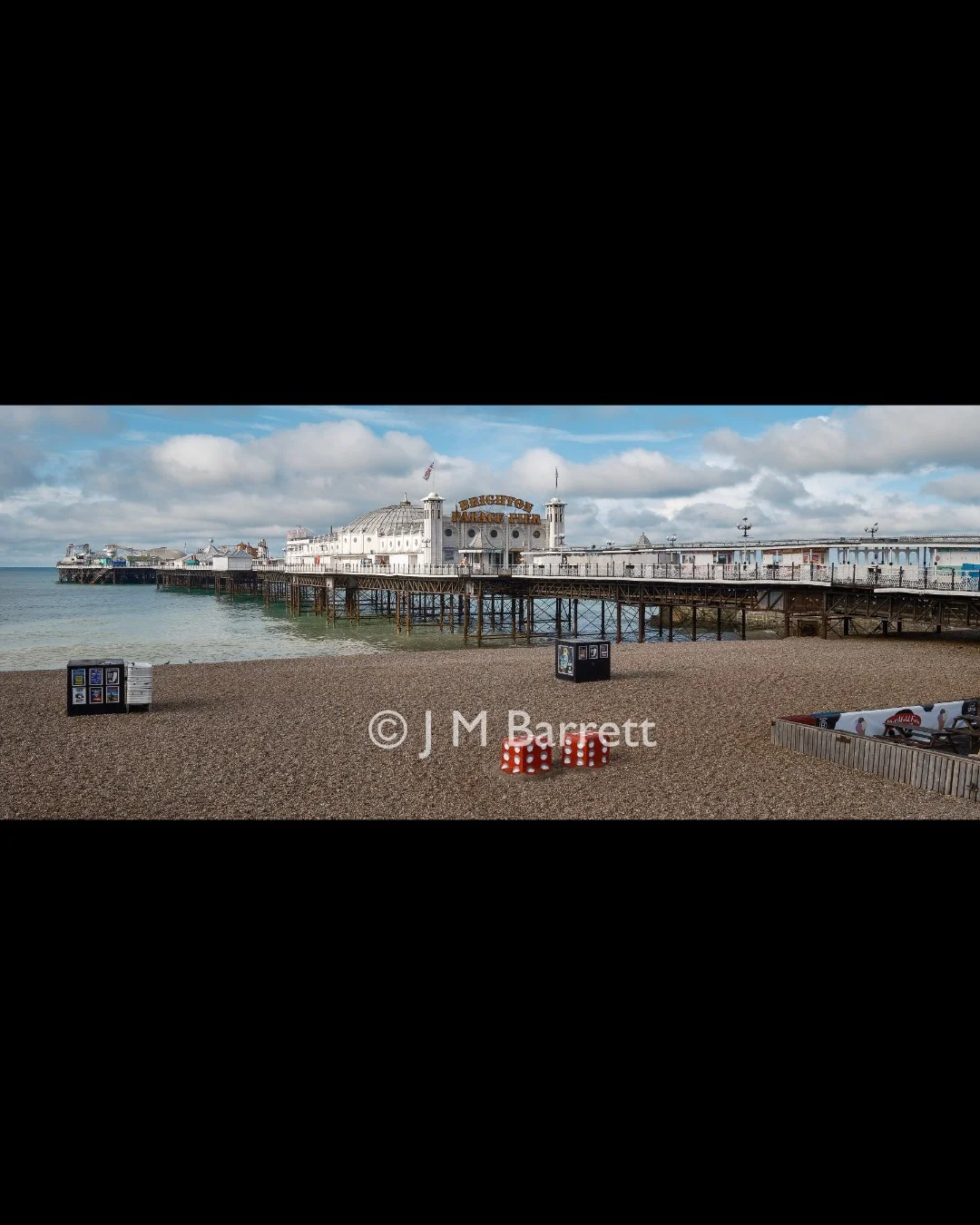 Brighton Pier extending into the ocean with a cloudy sky, and beach objects like dice and trash bins in the foreground.