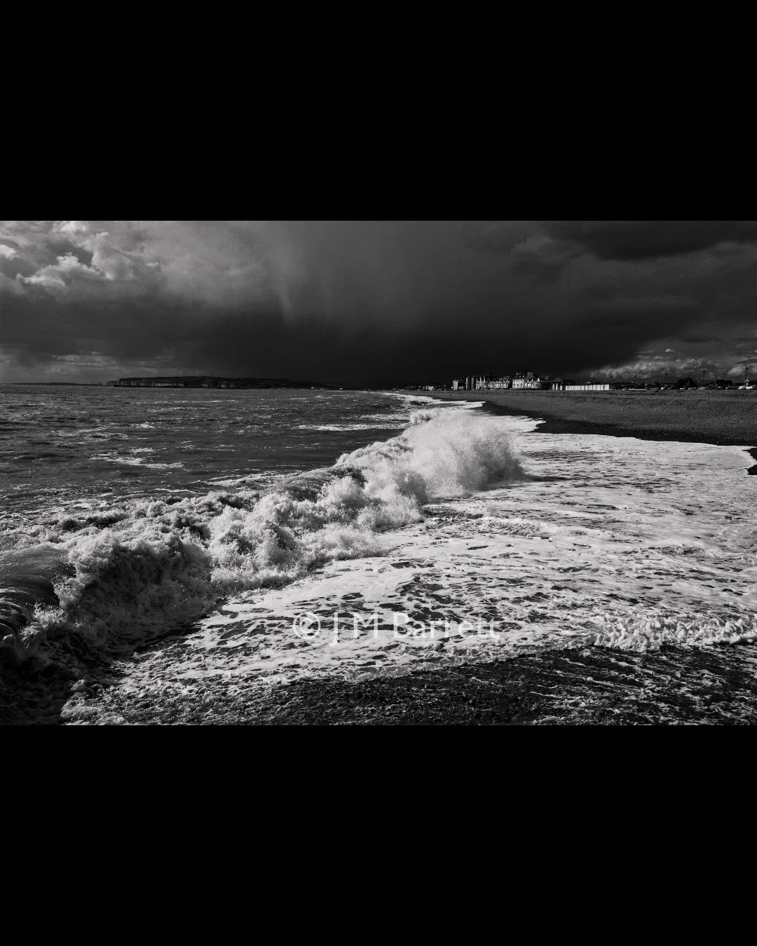Black and white photo of a beach with waves hitting the shore and dark storm clouds looming overhead.