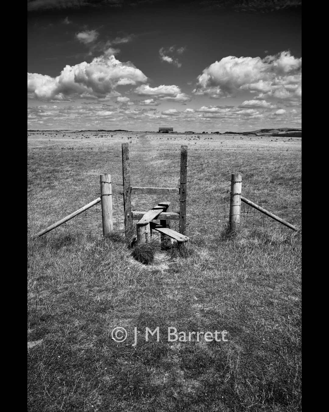 A black and white photo of a rural landscape with a barbed wire fence and a broken wooden gate in the foreground, an open field with scattered bushes, and a house or barn in the distance under a partly cloudy sky.