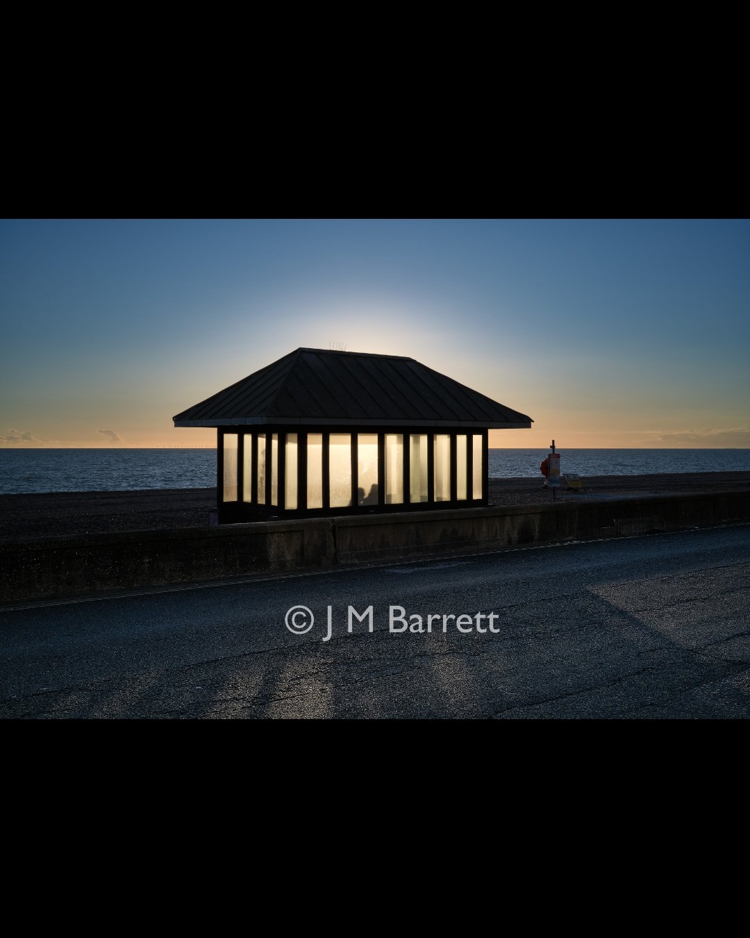 Seaside shelter at sunset with silhouette of person inside, ocean in the background, and a paved walkway in the foreground.