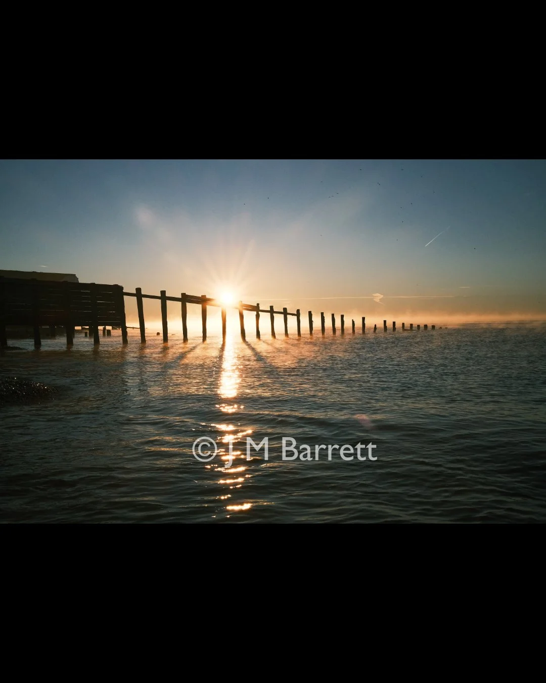 Sunrise over a wooden pier extending into the ocean with reflections on the water and a clear sky.
