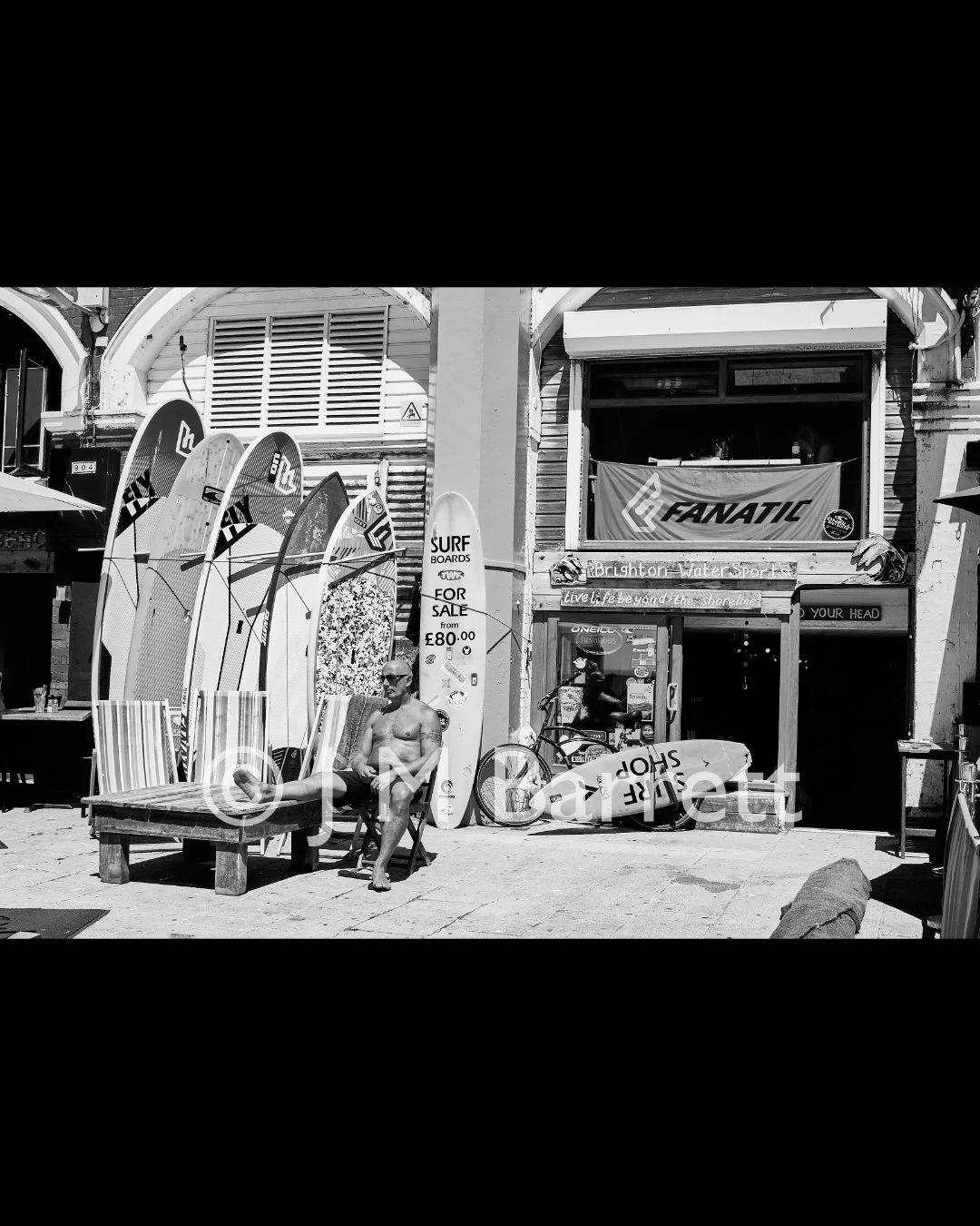 A man sitting shirtless on a chair outside a surf shop with surfboards for sale, a banner, and storefront signage on a busy street in Brighton, UK.