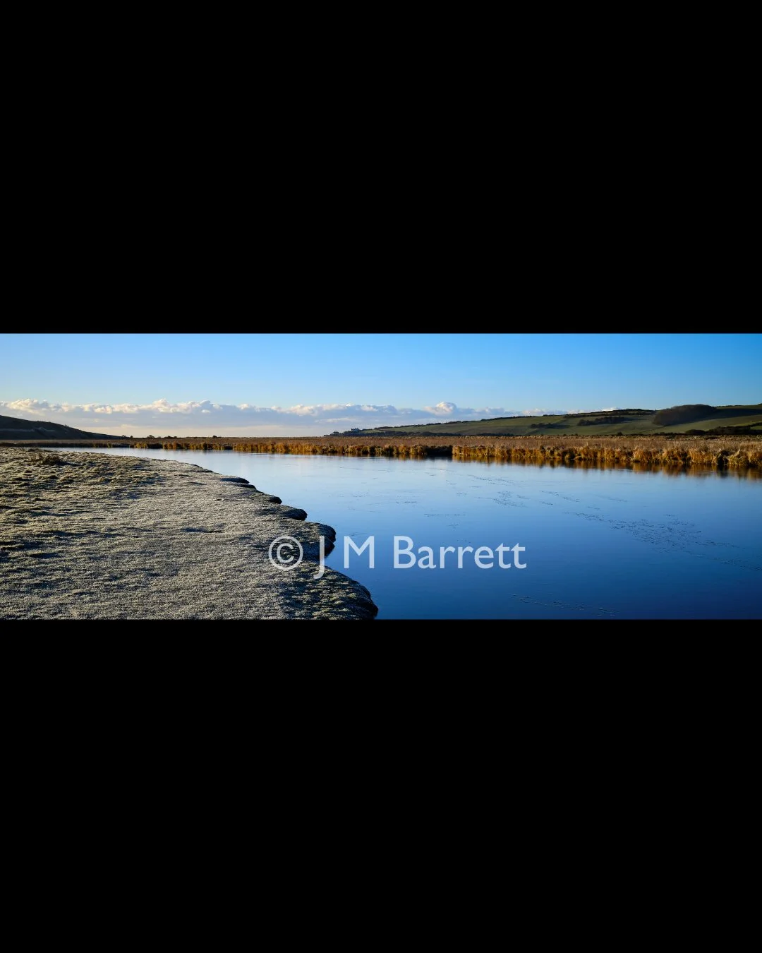 A calm river reflecting the blue sky and scattered clouds, with grassy banks and rolling hills in the background.